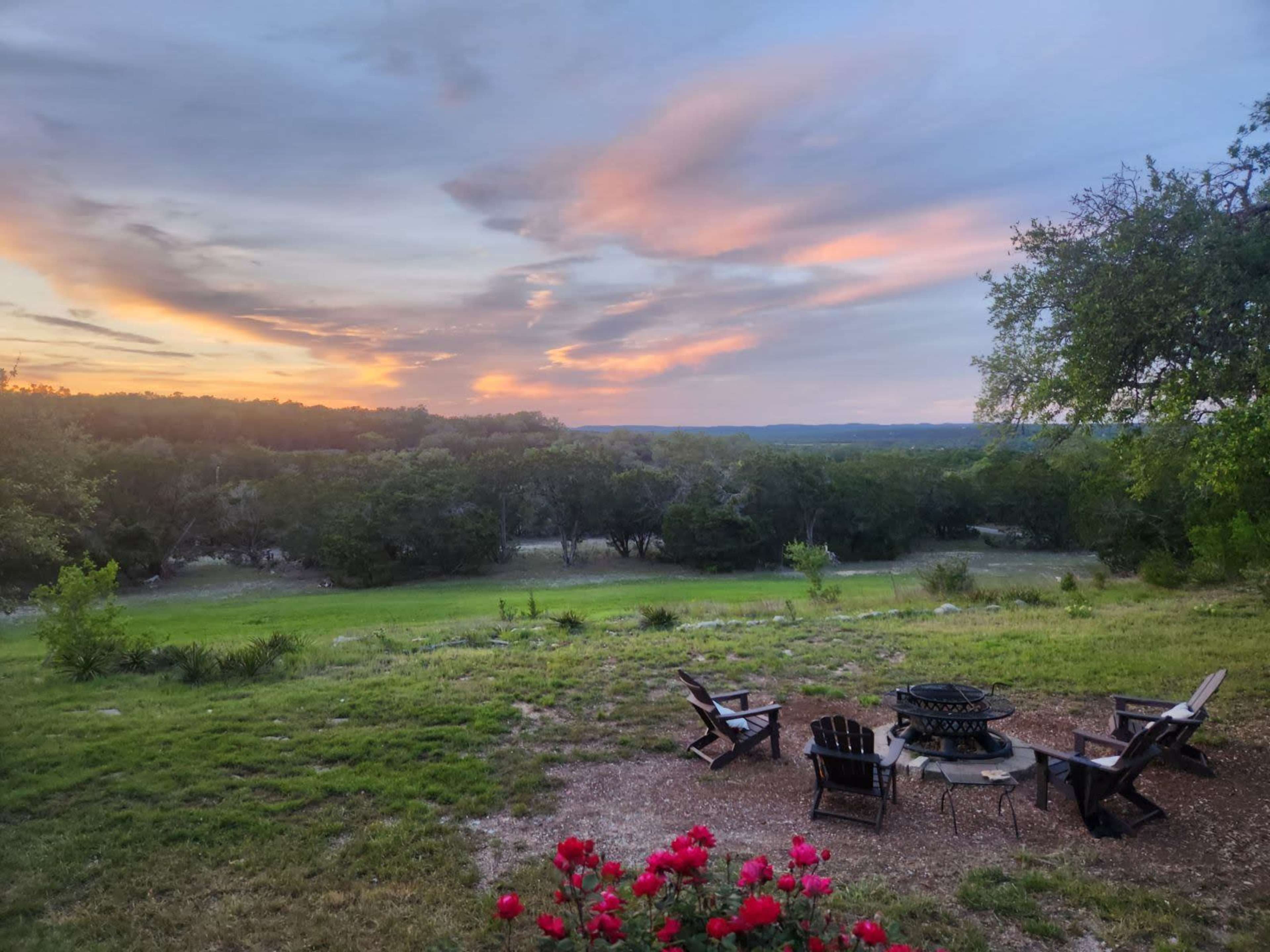 A fire pit surrounded by Adirondack chairs overlooks a grassy area and trees at sunset.