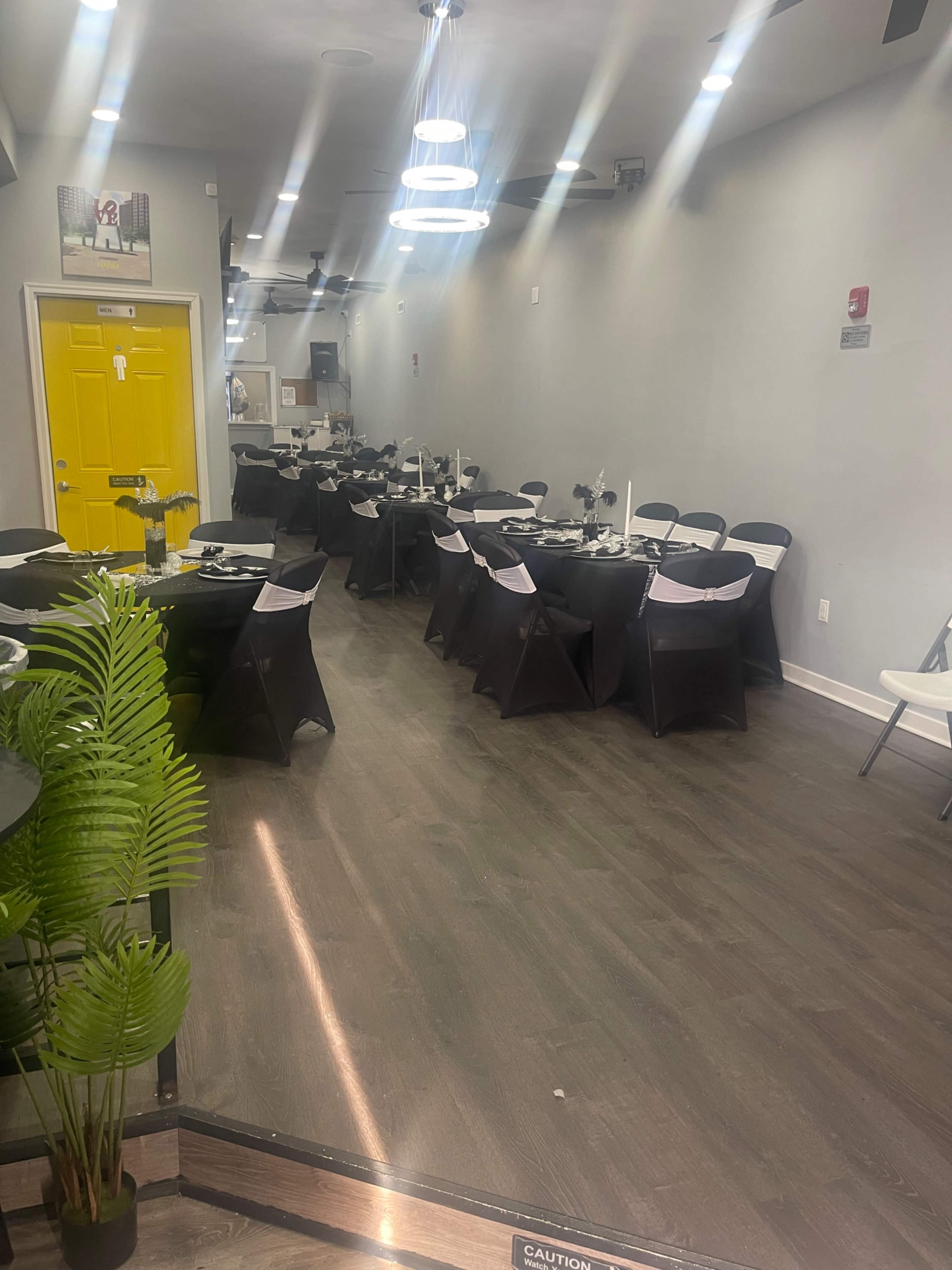 The image shows an indoor dining area arranged with black tables and chairs, featuring a yellow door in the background.