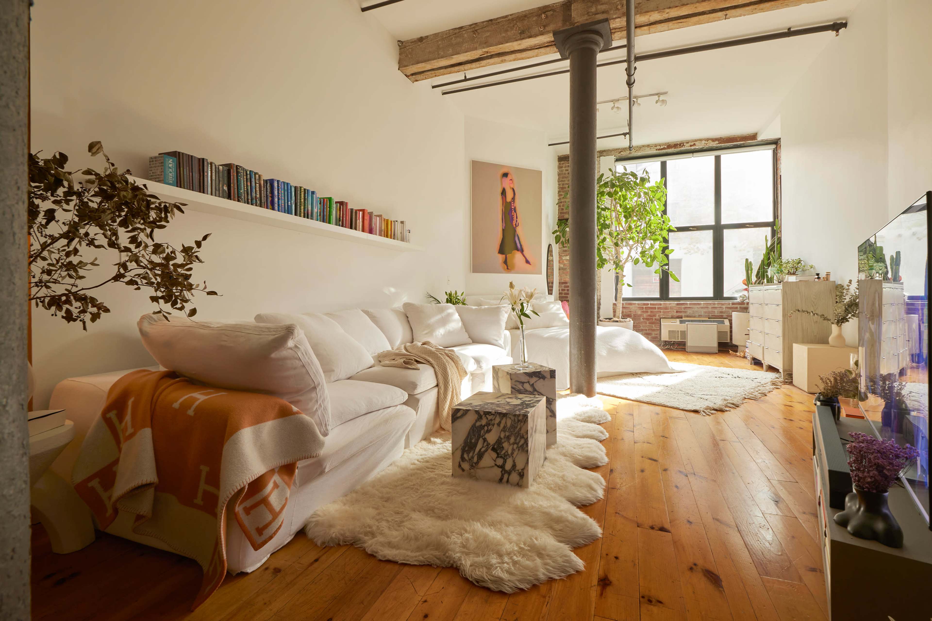 The image shows a bright living room with a white sectional sofa, wooden flooring, plants, and a large window letting in natural light.