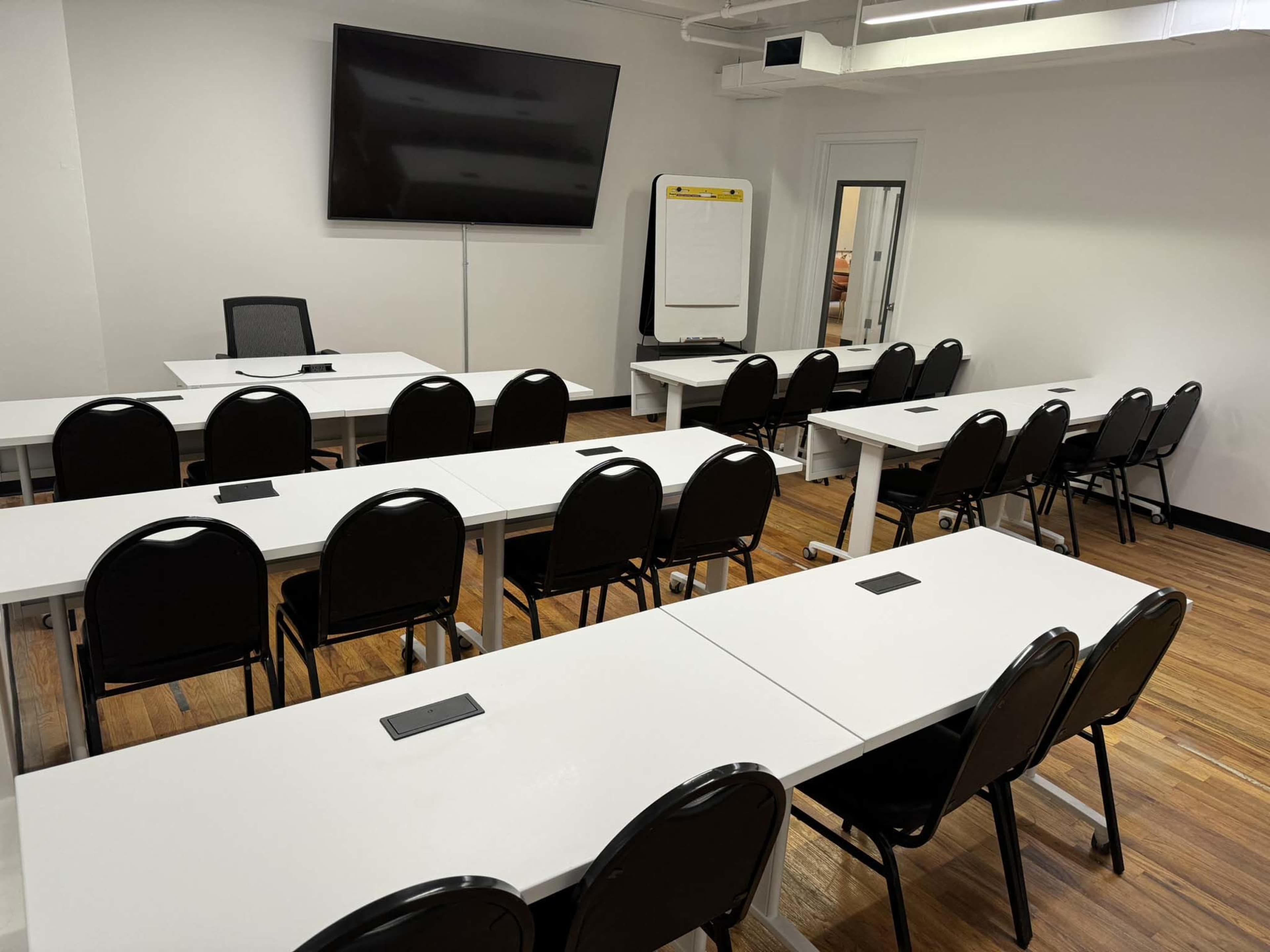 A classroom setup with several rows of tables and chairs, a large wall-mounted TV, and a flip chart in the corner.