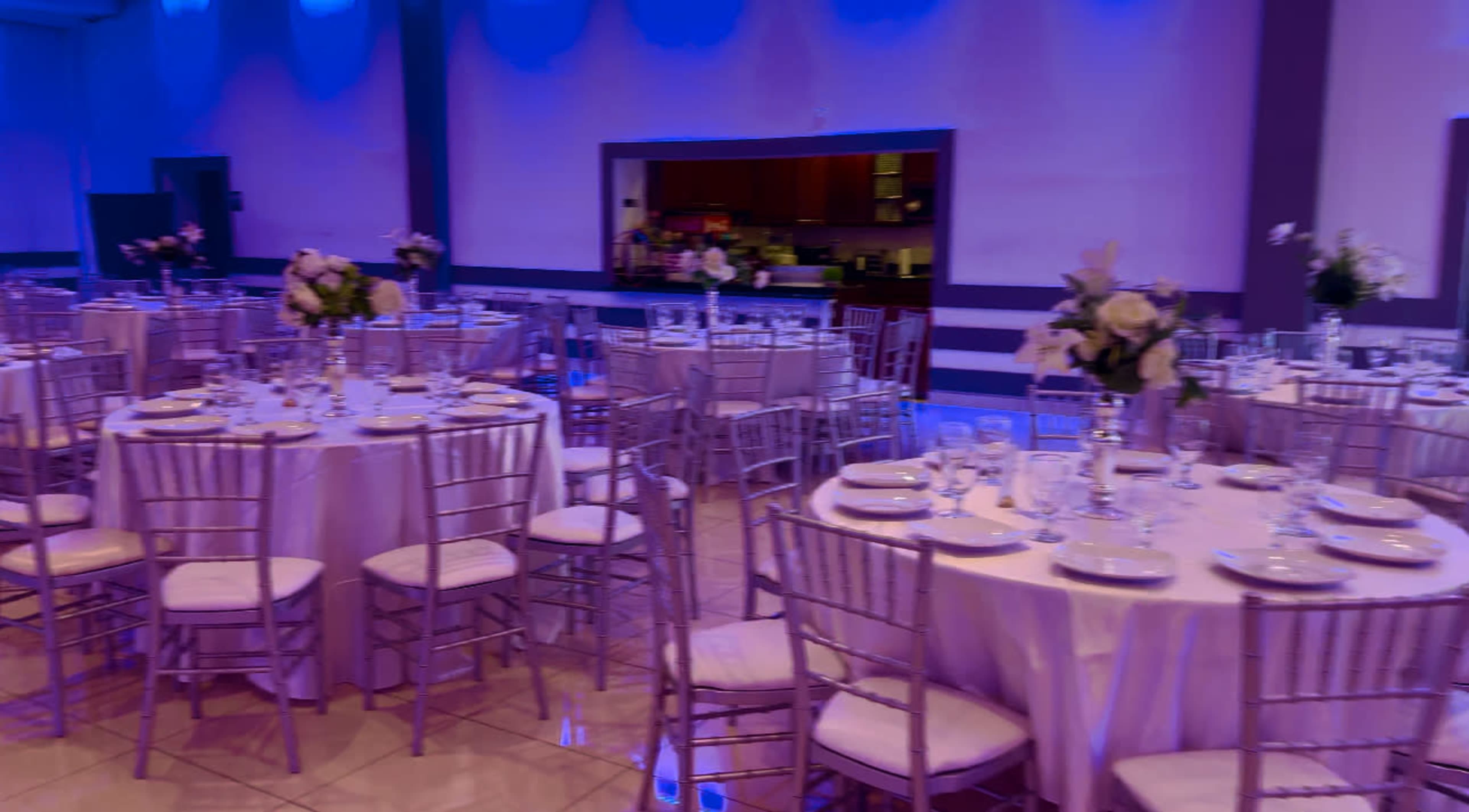 A banquet hall features round tables set with dishes and floral centerpieces under blue ambient lighting.