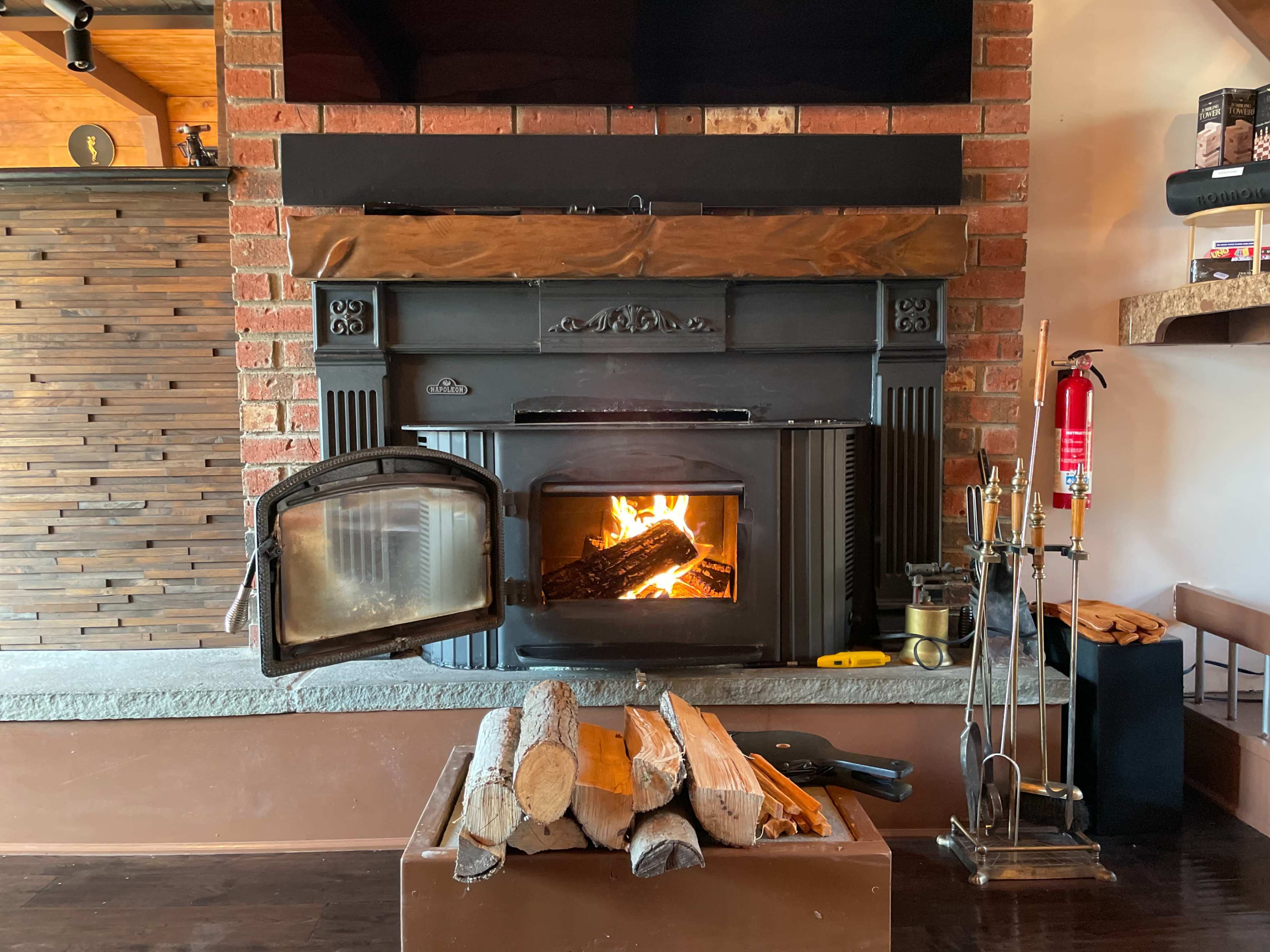 A wood-burning stove with an open door, surrounded by stacked logs and various fireplace tools on a wooden floor.