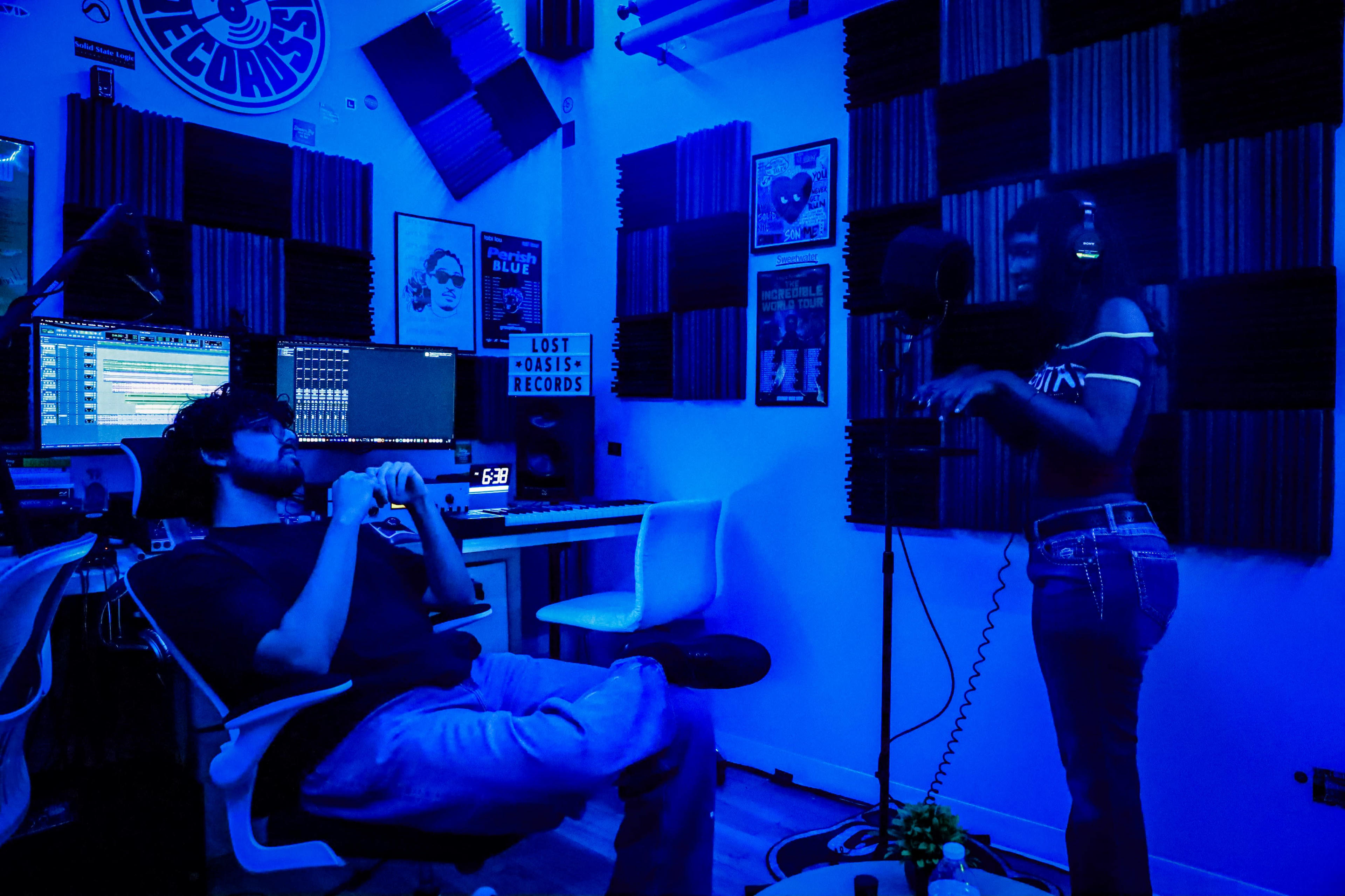 A woman stands in front of a microphone in a recording studio while a man sits in a chair, observing her performance.
