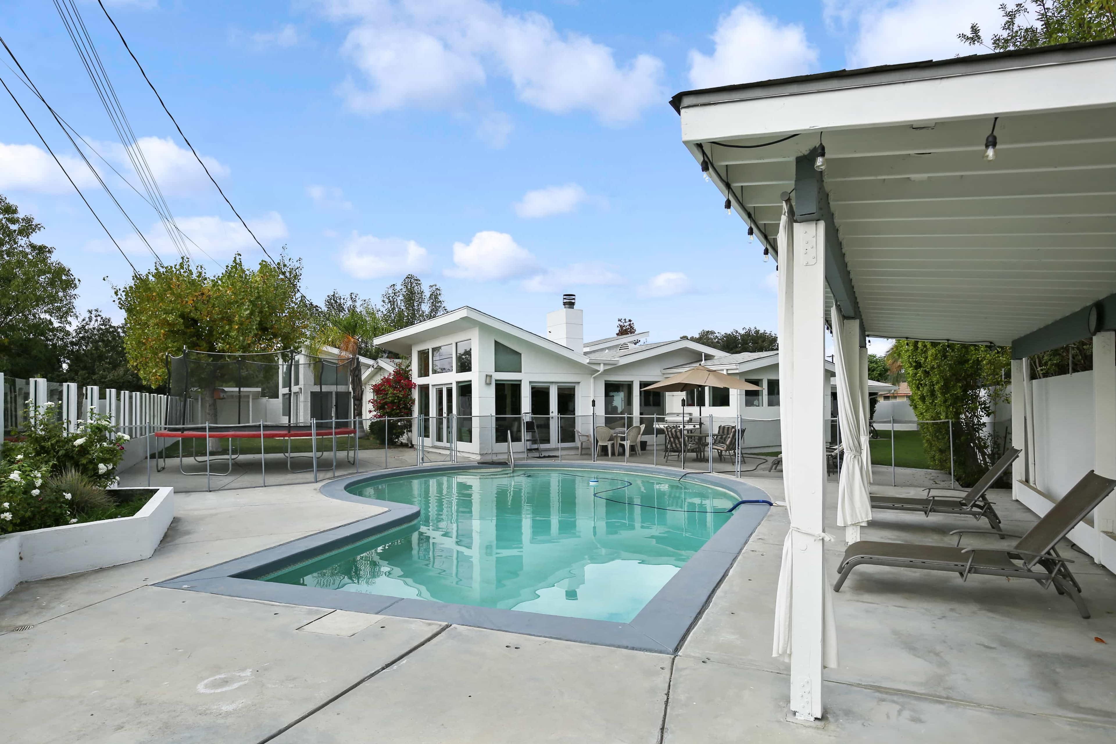 A backyard with a swimming pool, surrounded by a patio area and a trampoline, alongside a modern-style house.
