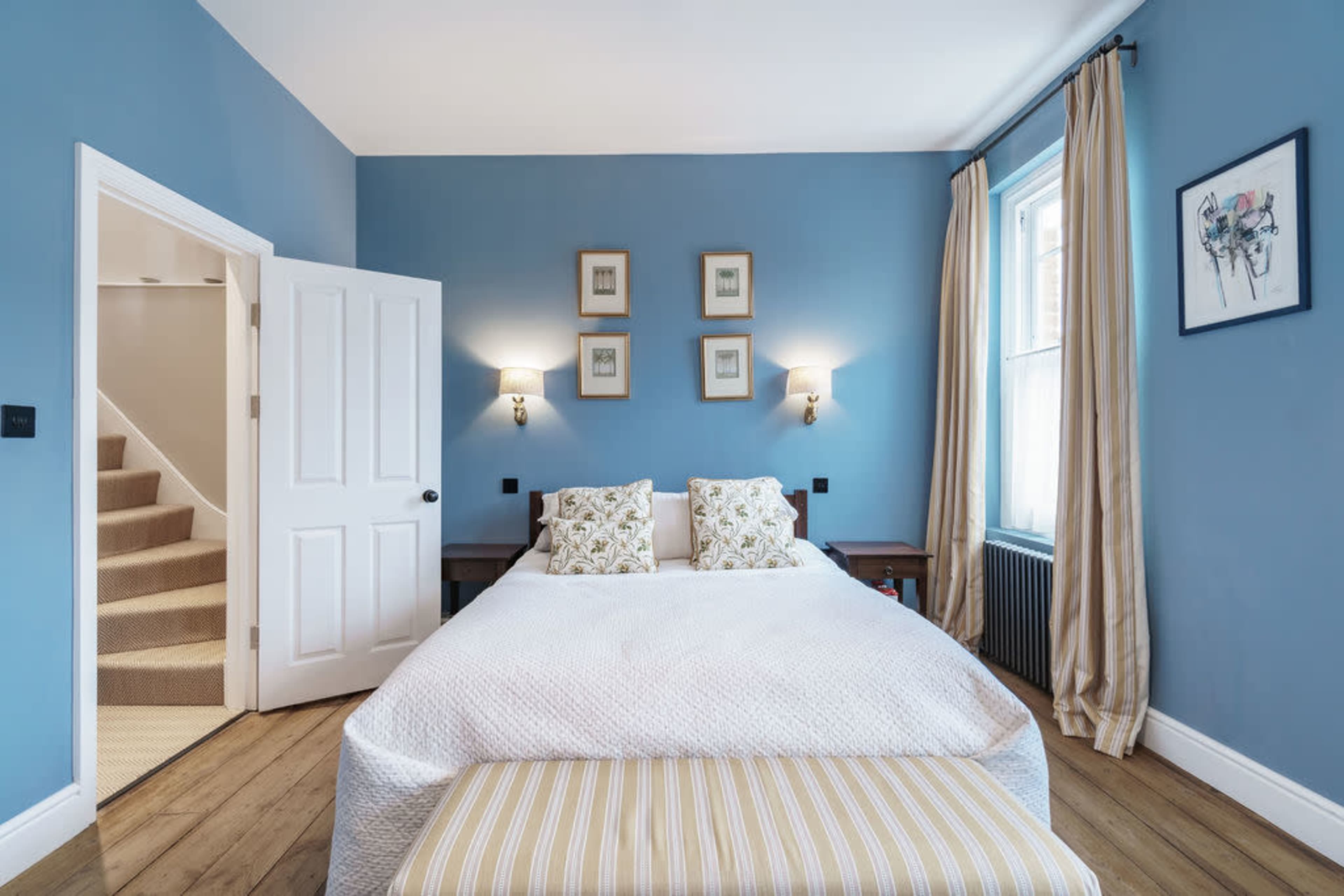 A bedroom with a blue wall, a double bed with decorative pillows, bedside lamps, and a staircase visible through an open door.