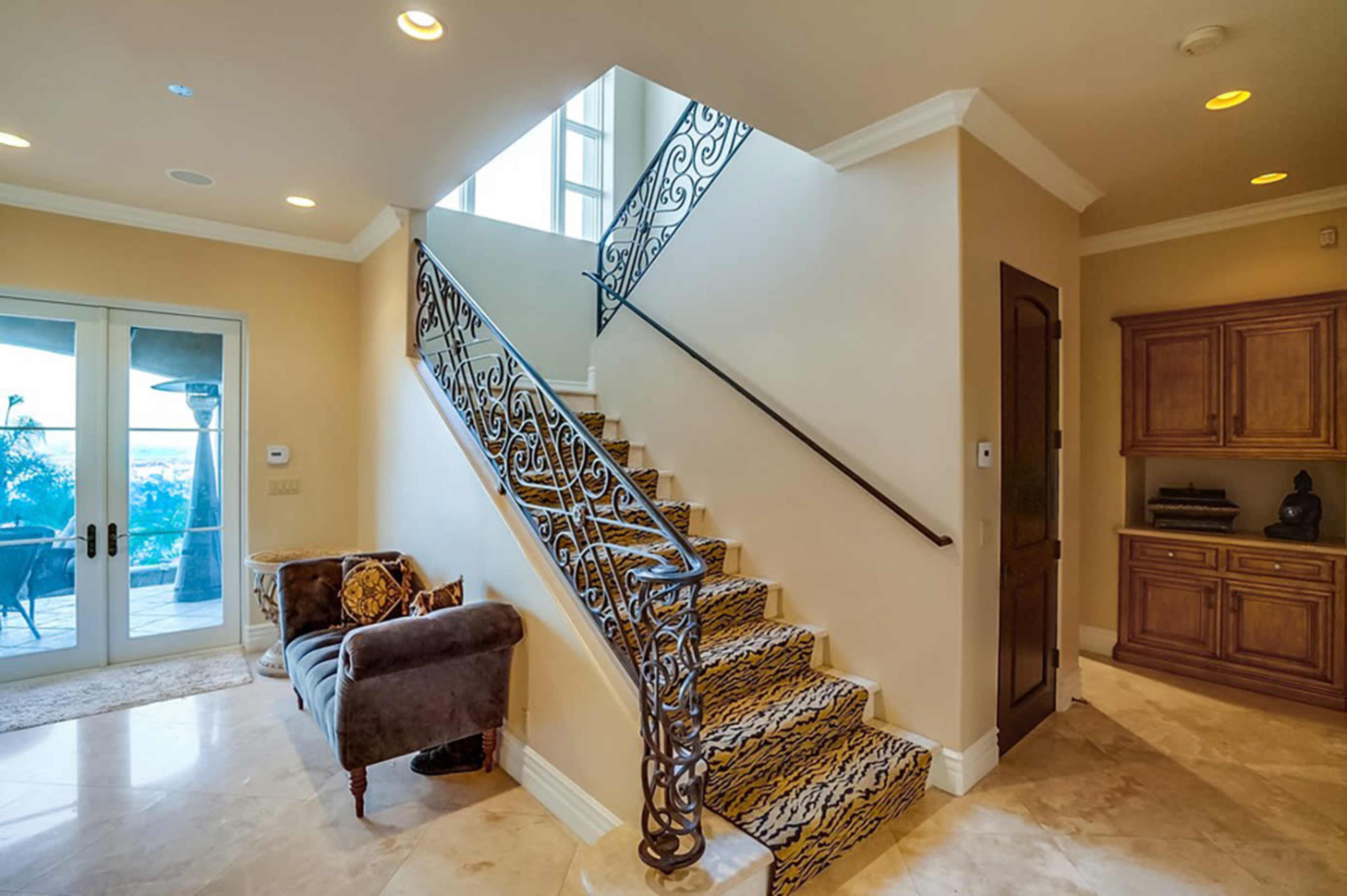 A staircase with ornate wrought iron railings leads to a second floor in a spacious foyer with marble flooring and a view of the outdoors through large windows.