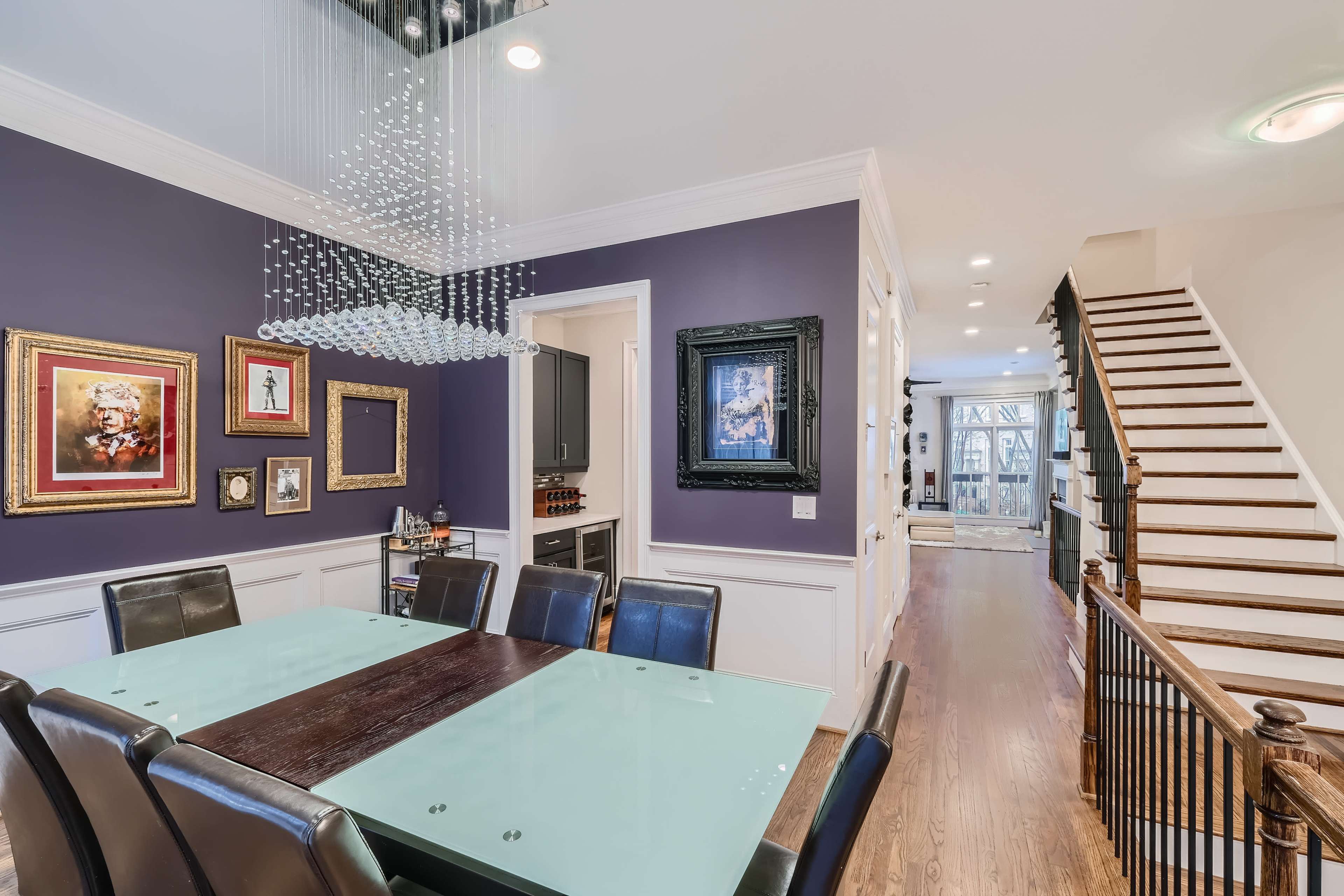 The image shows a modern dining room with a glass-topped table surrounded by black chairs, framed artwork on the purple walls, and a staircase leading to another level.