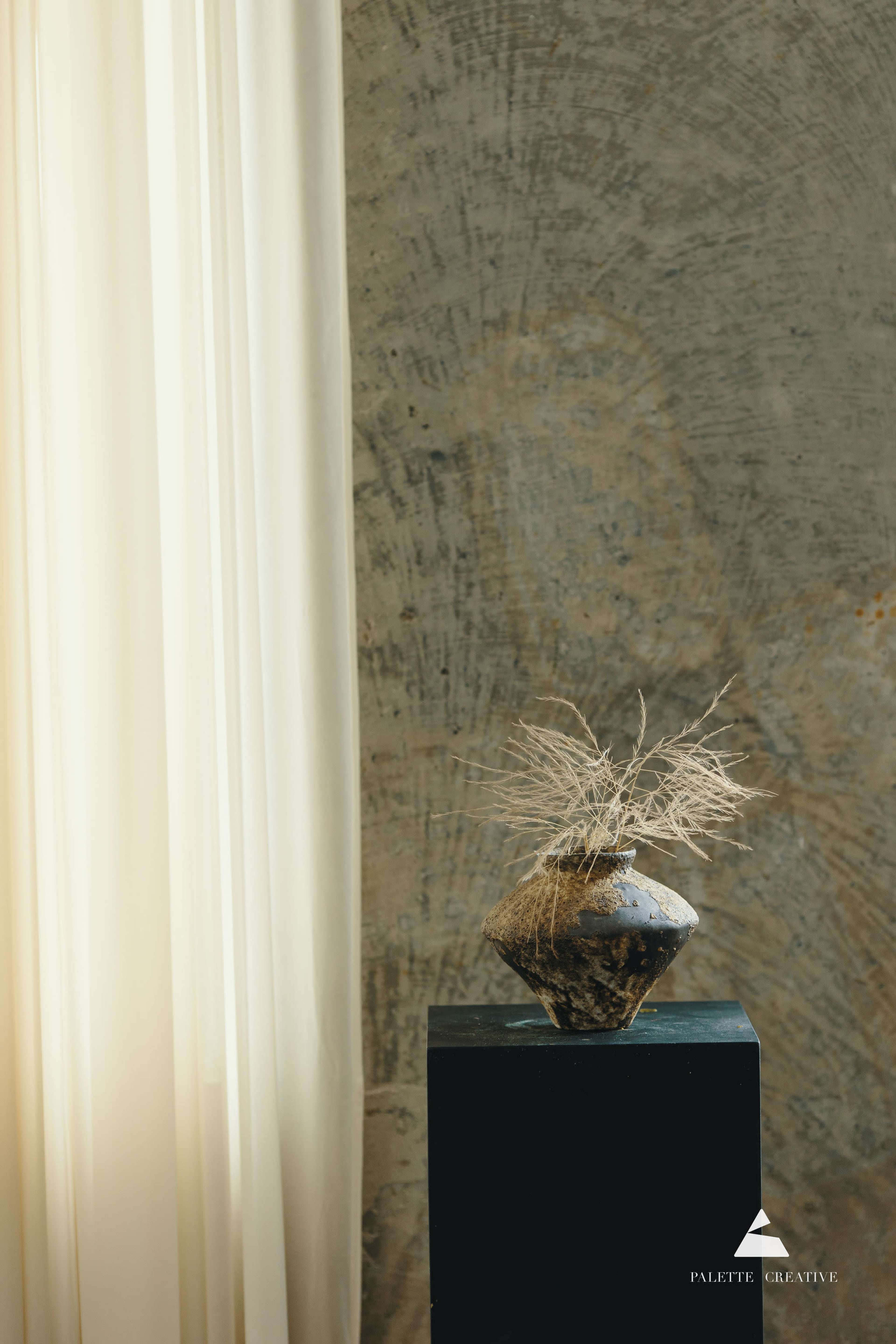A dark, textured vase with dried plant material sits atop a black pedestal beside a sheer, light-colored curtain and a concrete wall.