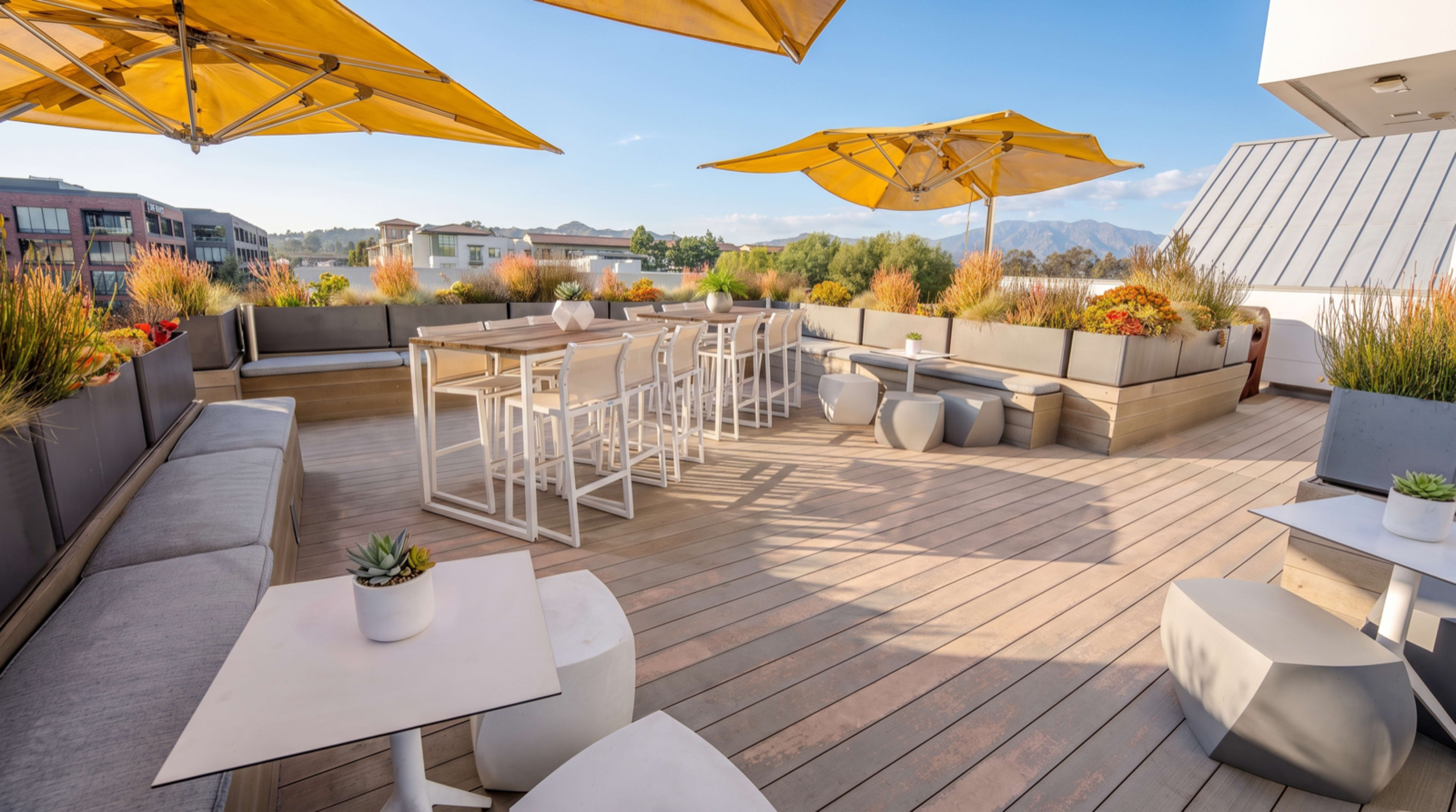 The image shows a rooftop terrace featuring wooden decking, furniture for dining and lounging, and yellow umbrellas providing shade.
