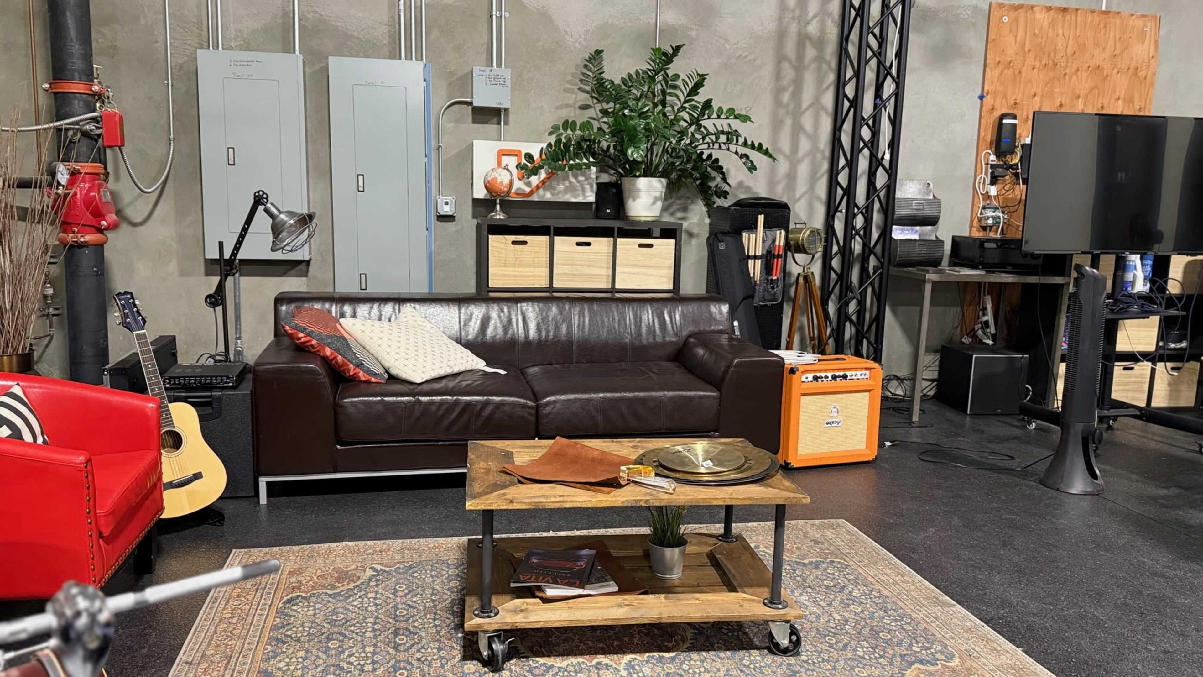 The image shows a modern, industrial-style studio setup with a brown leather sofa, a wooden coffee table, a guitar, and various functional furniture including storage cabinets and a flat-screen TV.