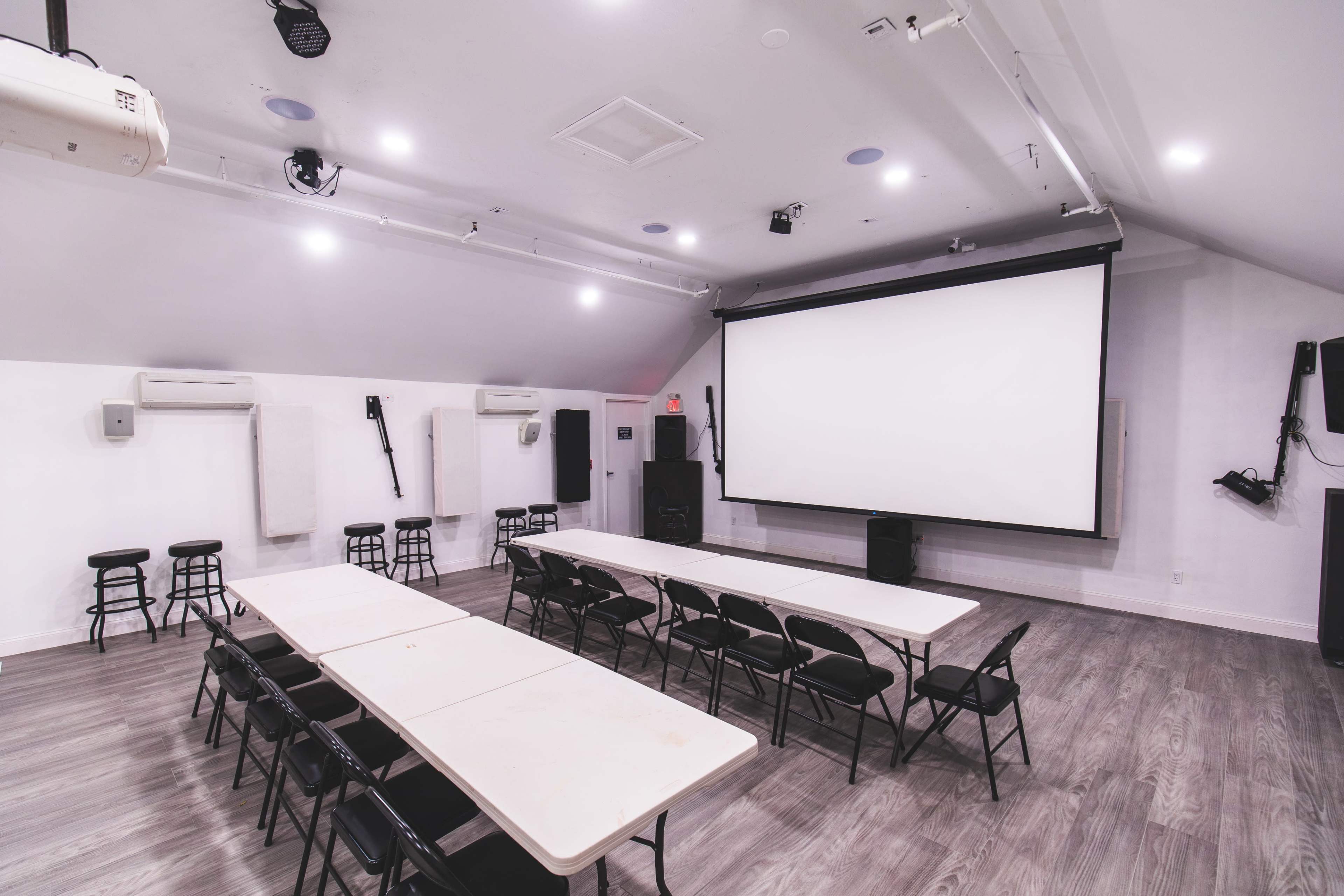 The image shows a spacious room set up for a presentation, featuring tables arranged in a U-shape, a large screen at the front, and seating along the sides.