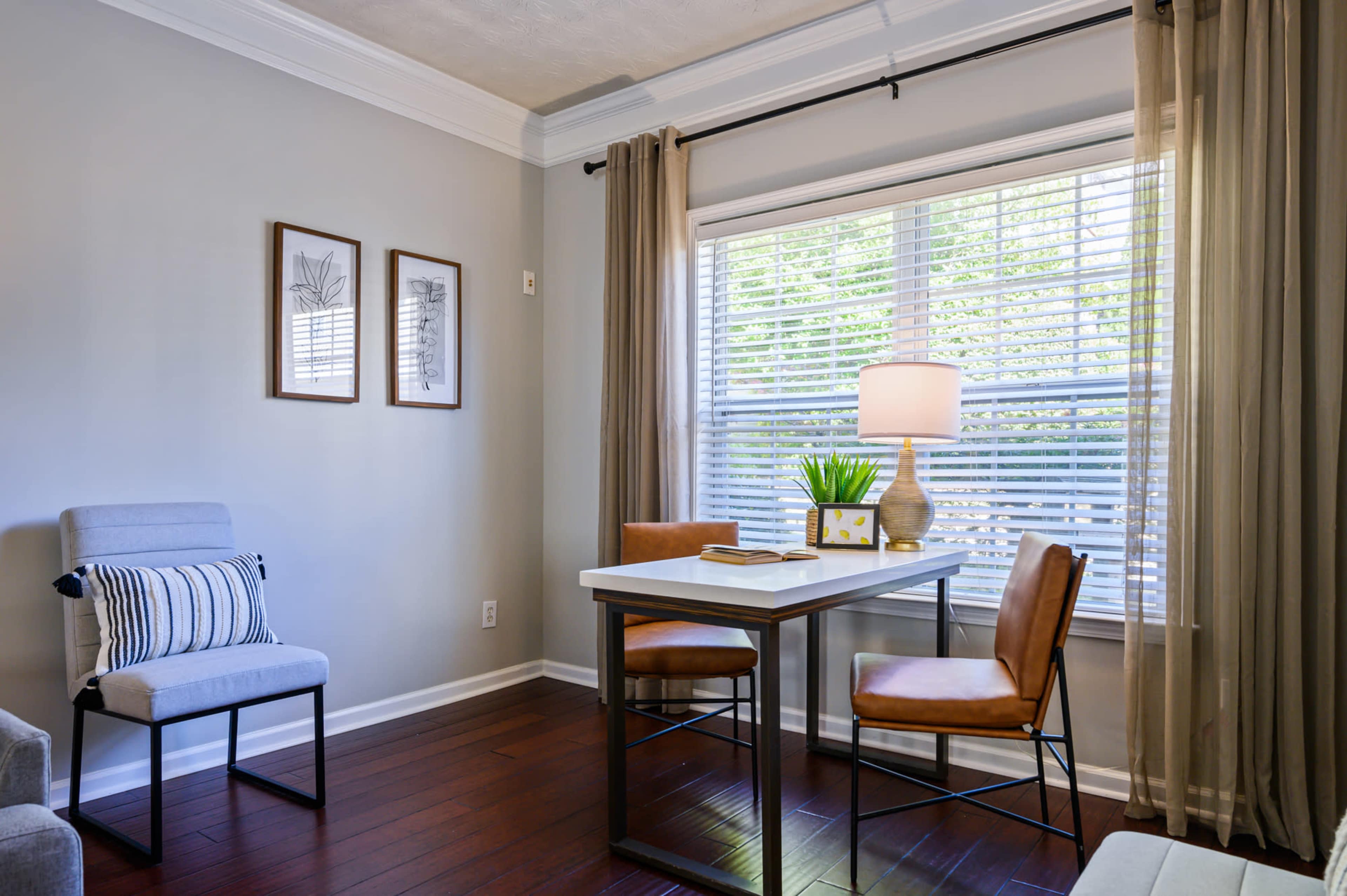 A small dining area features a table with two chairs, a lamp, and decorative items beside a window with blinds.