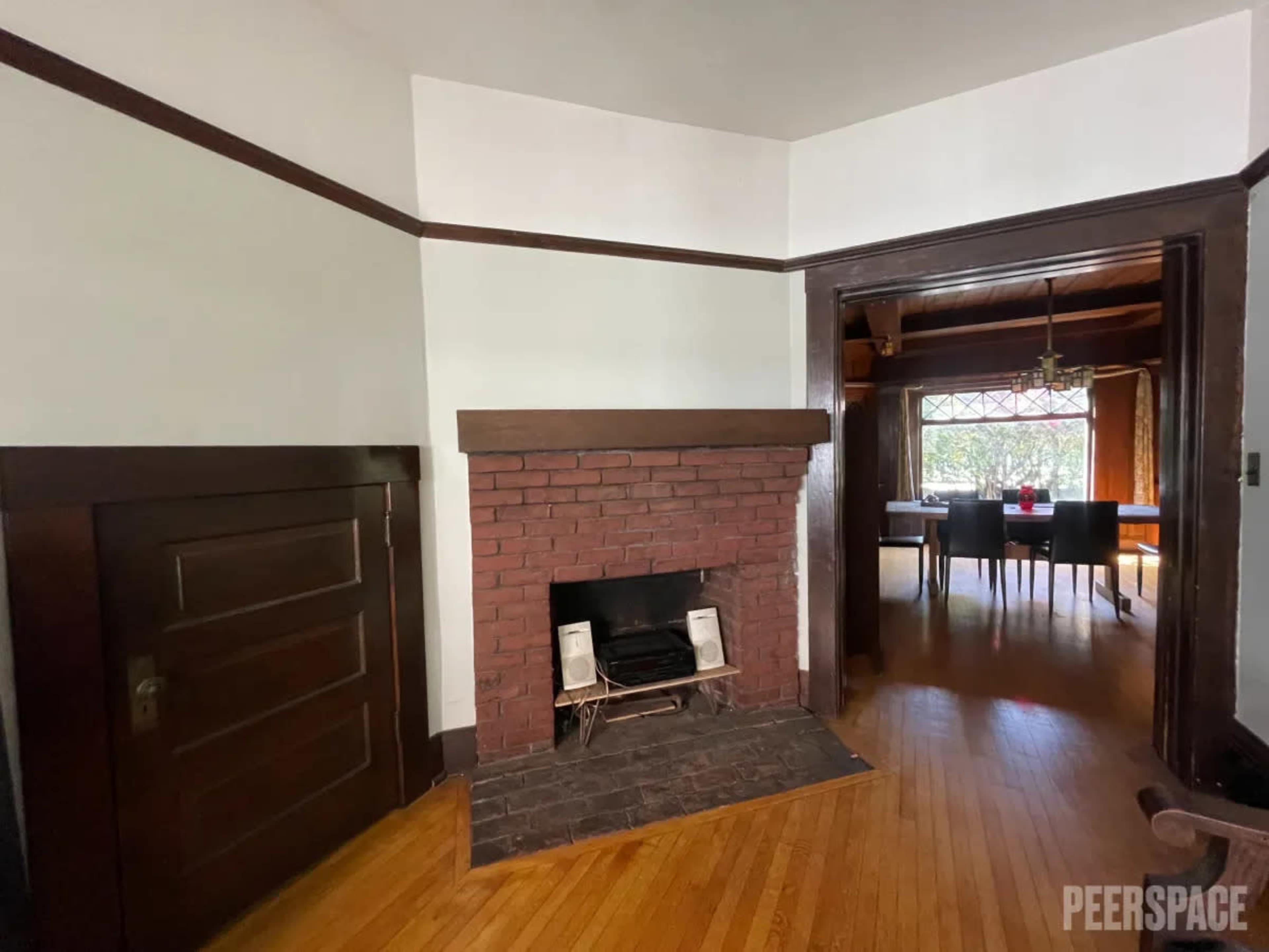 A brick fireplace with a dark wooden mantel, adjacent to a doorway leading to a room with a dining area visible through the opening.