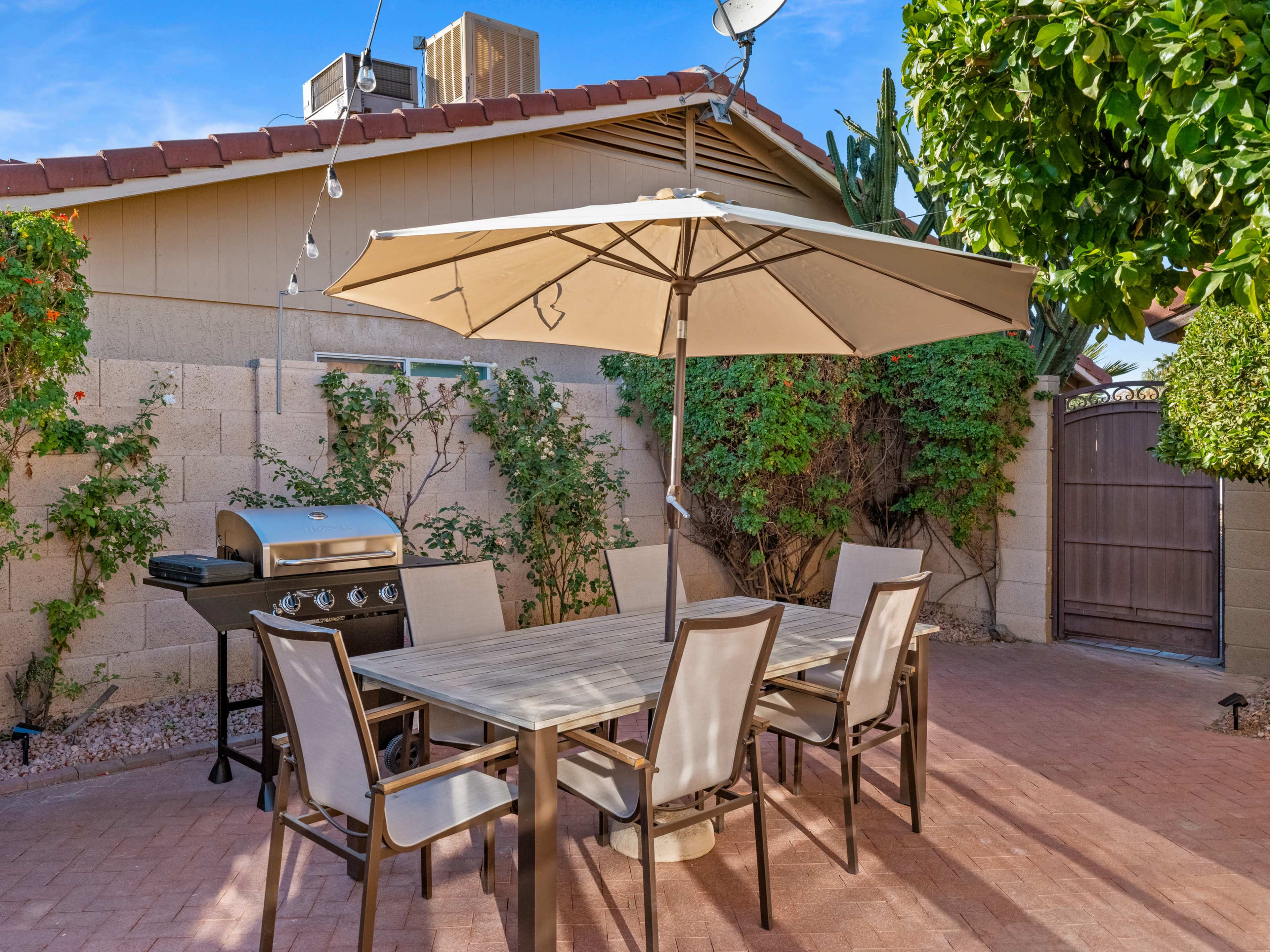 A patio features a large umbrella over a dining table surrounded by chairs, with a barbecue grill and greenery in the background.