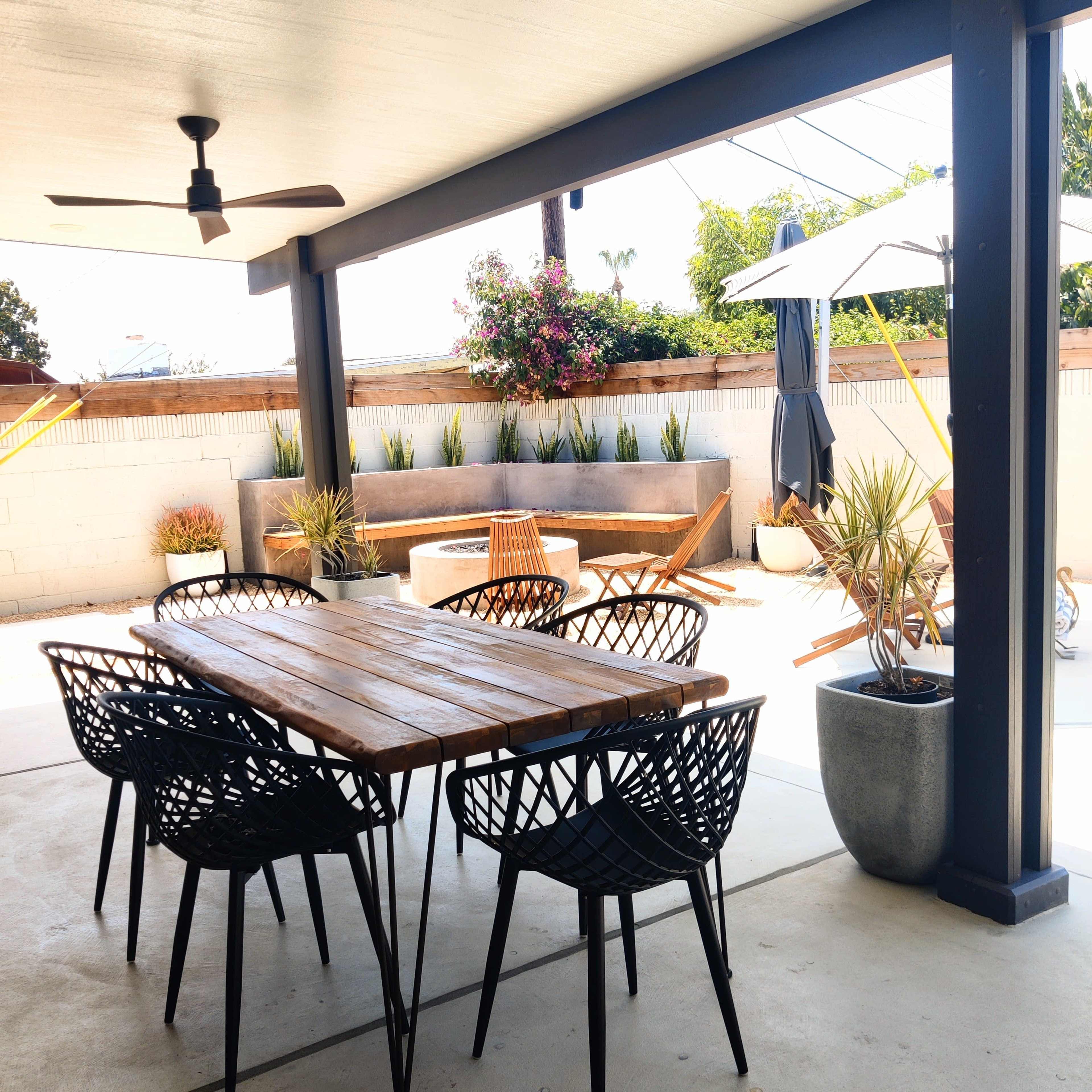 A wooden dining table surrounded by black chairs is positioned under a covered patio, with a landscaped outdoor area featuring lounge chairs, potted plants, and a sun umbrella in the background.