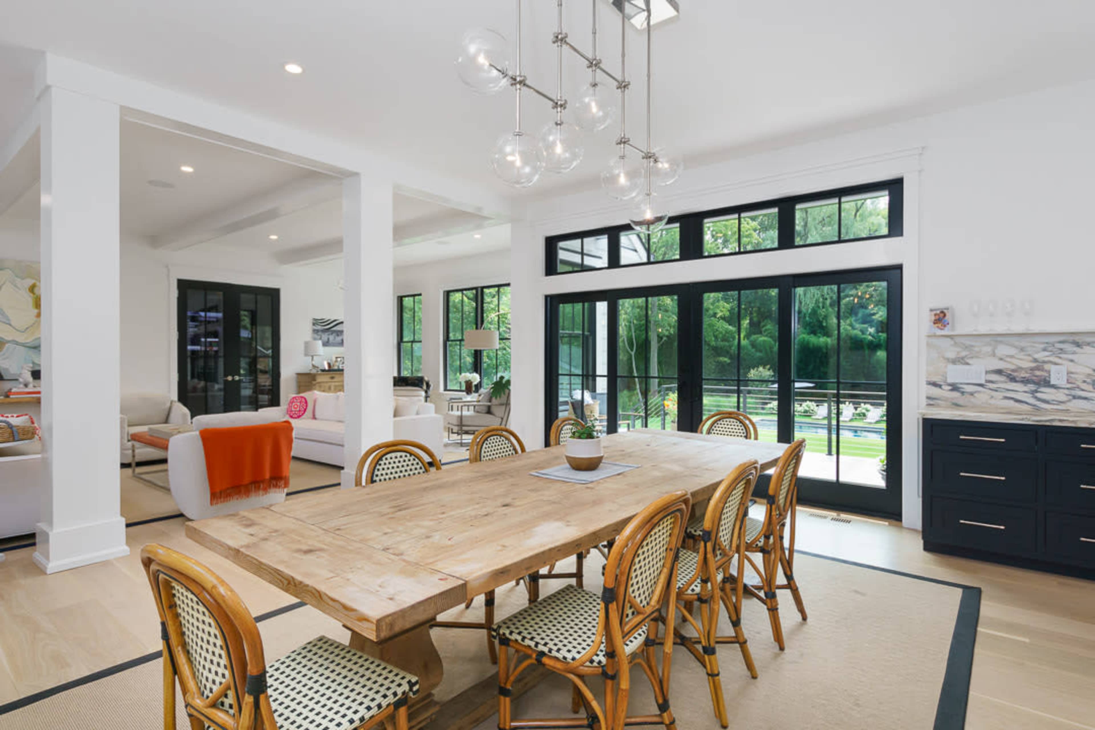 A long wooden dining table with rattan chairs is positioned in a bright open-concept dining area, adjacent to a modern kitchen and living space with large windows overlooking a green outdoor area.