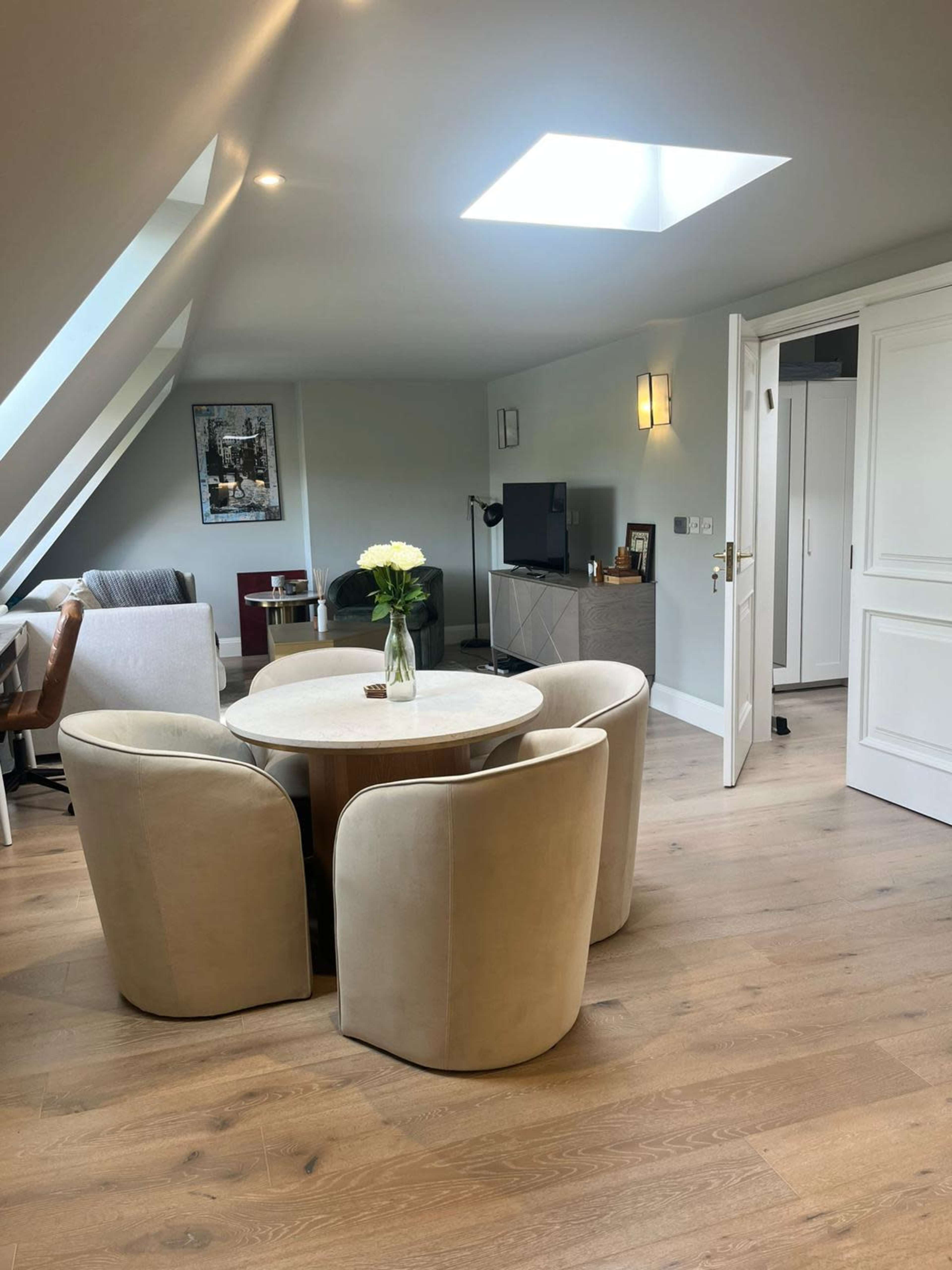 The image shows a modern, well-lit living space with a circular table and four upholstered chairs, a television unit, and a skylight.