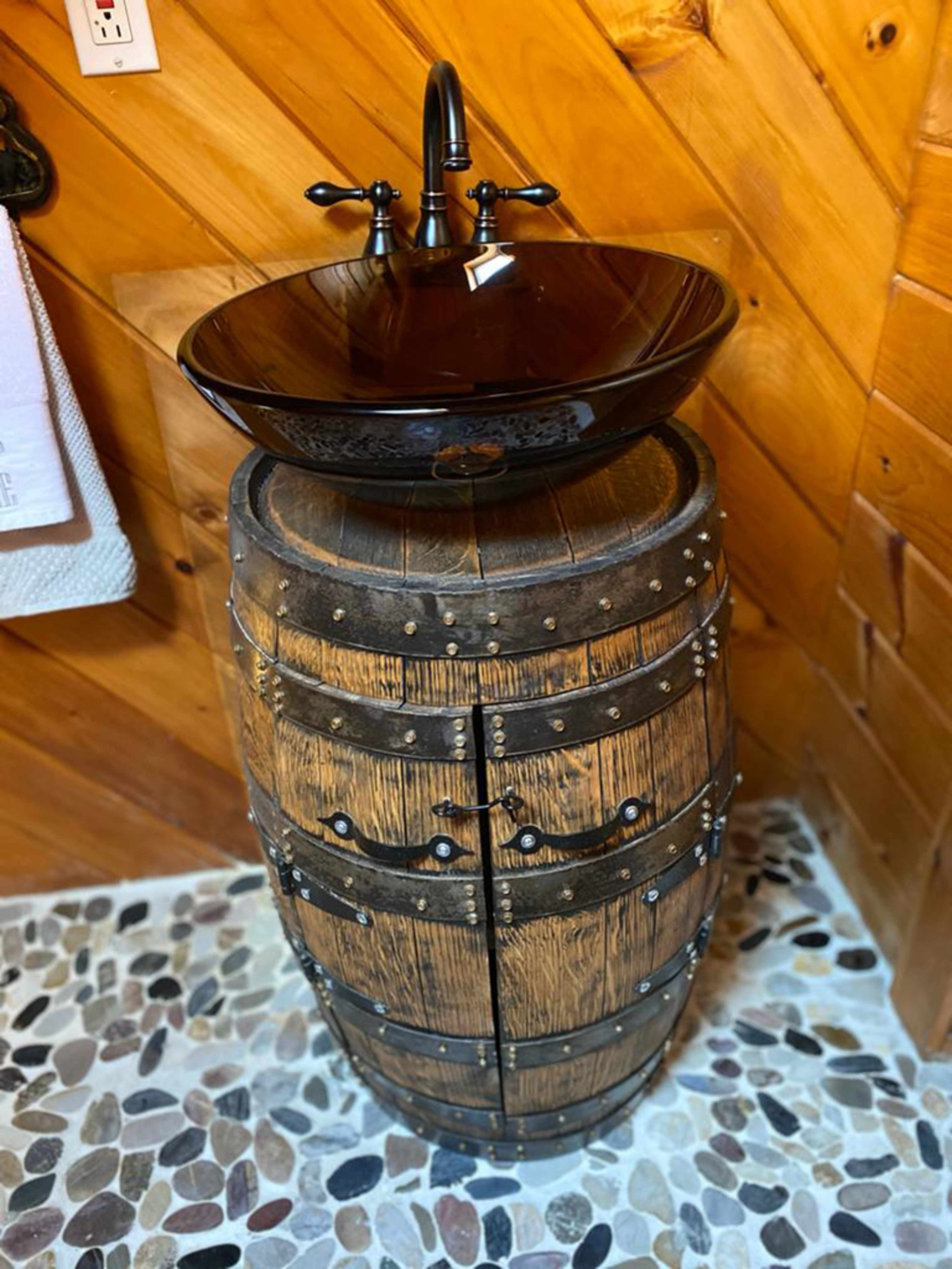 A black bowl sink set on a wooden barrel, positioned against a wooden wall with a pebble floor.