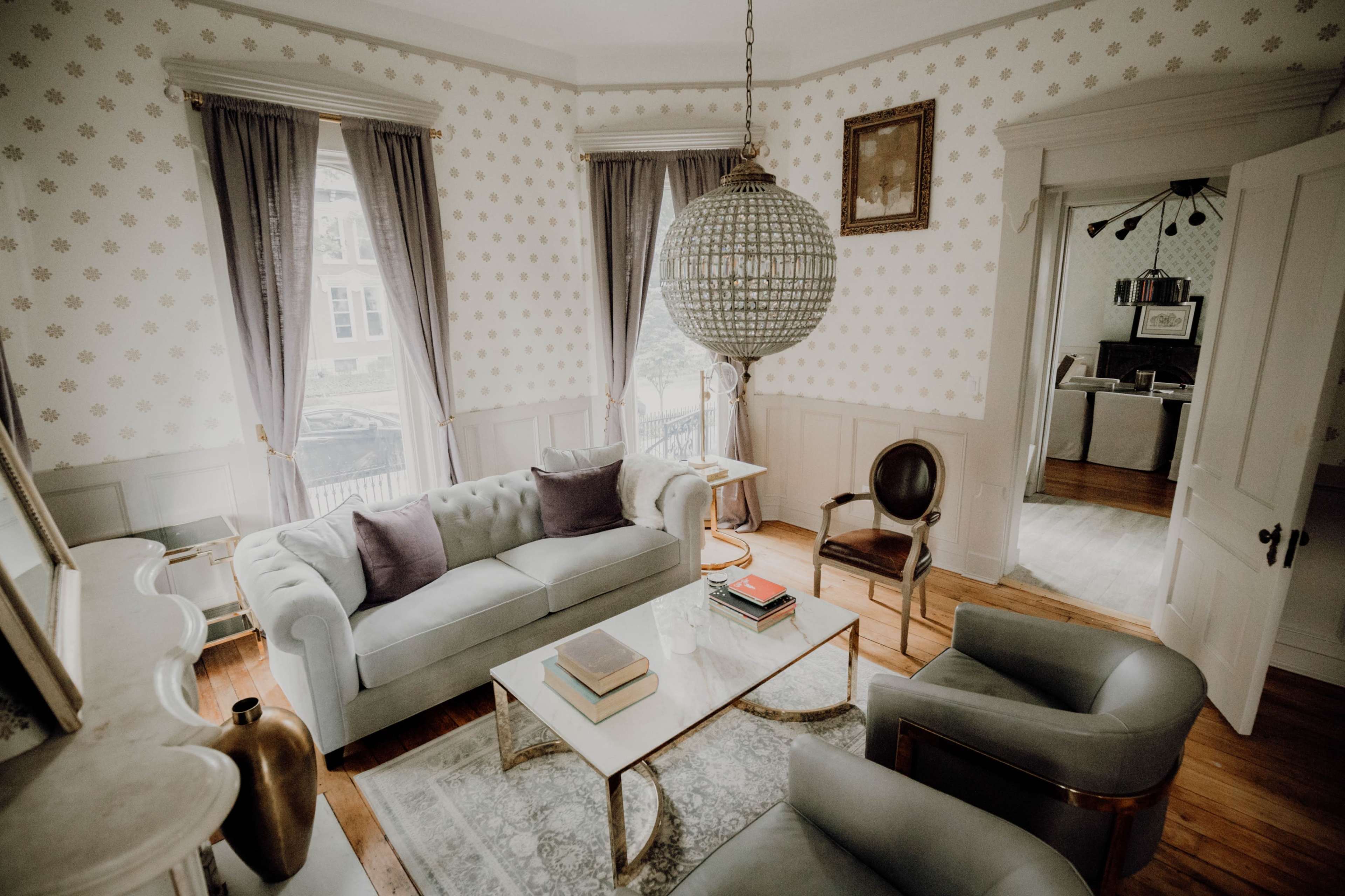 The room features a light-colored sofa with two dark cushions, a glass coffee table, and a large pendant light, with large windows dressed in curtains and a hardwood floor.