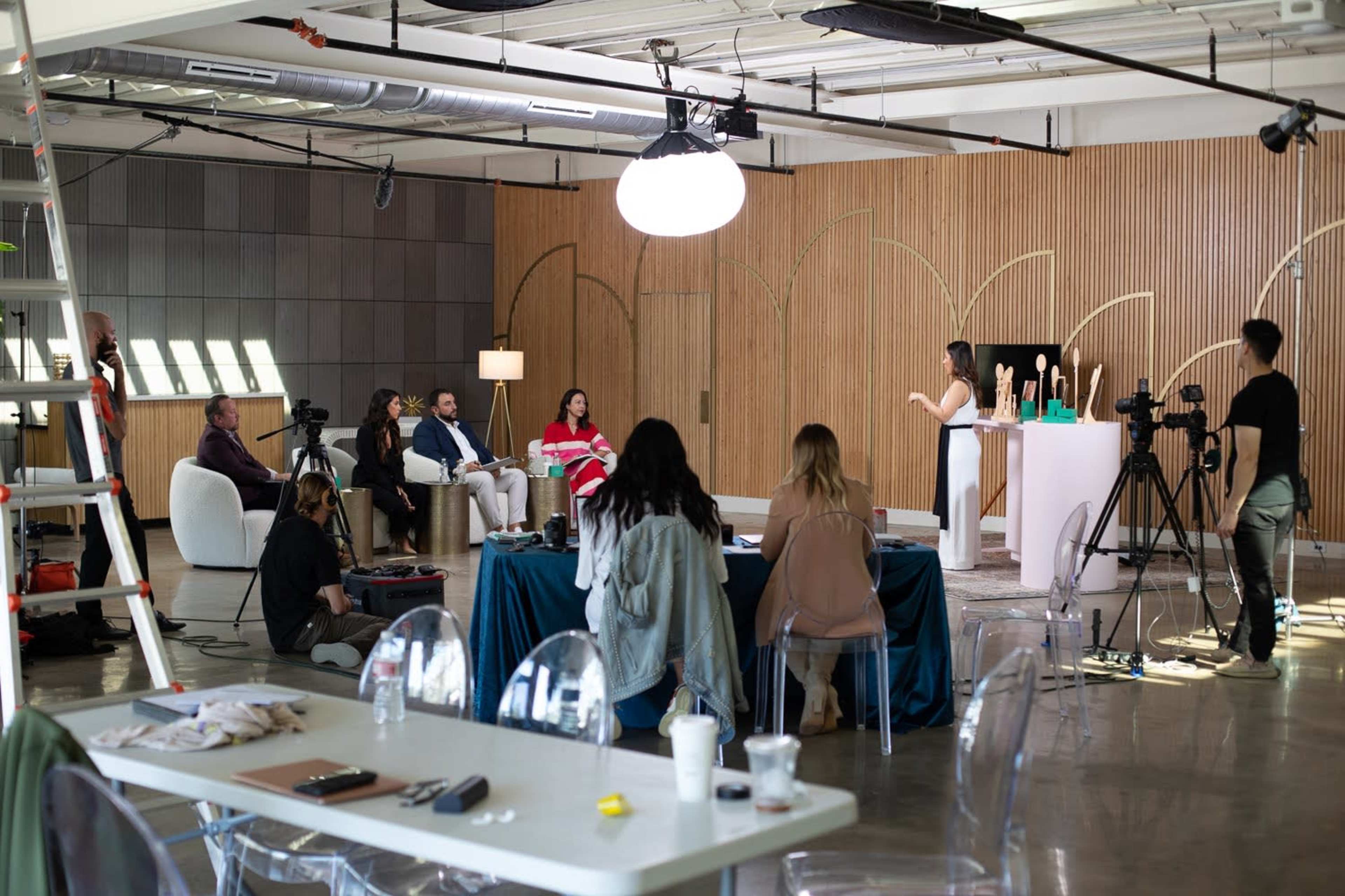 A group of people are seated in a studio, observing a presenter who is speaking in front of a wooden backdrop with sculptures, while cameras are set up to capture the event.