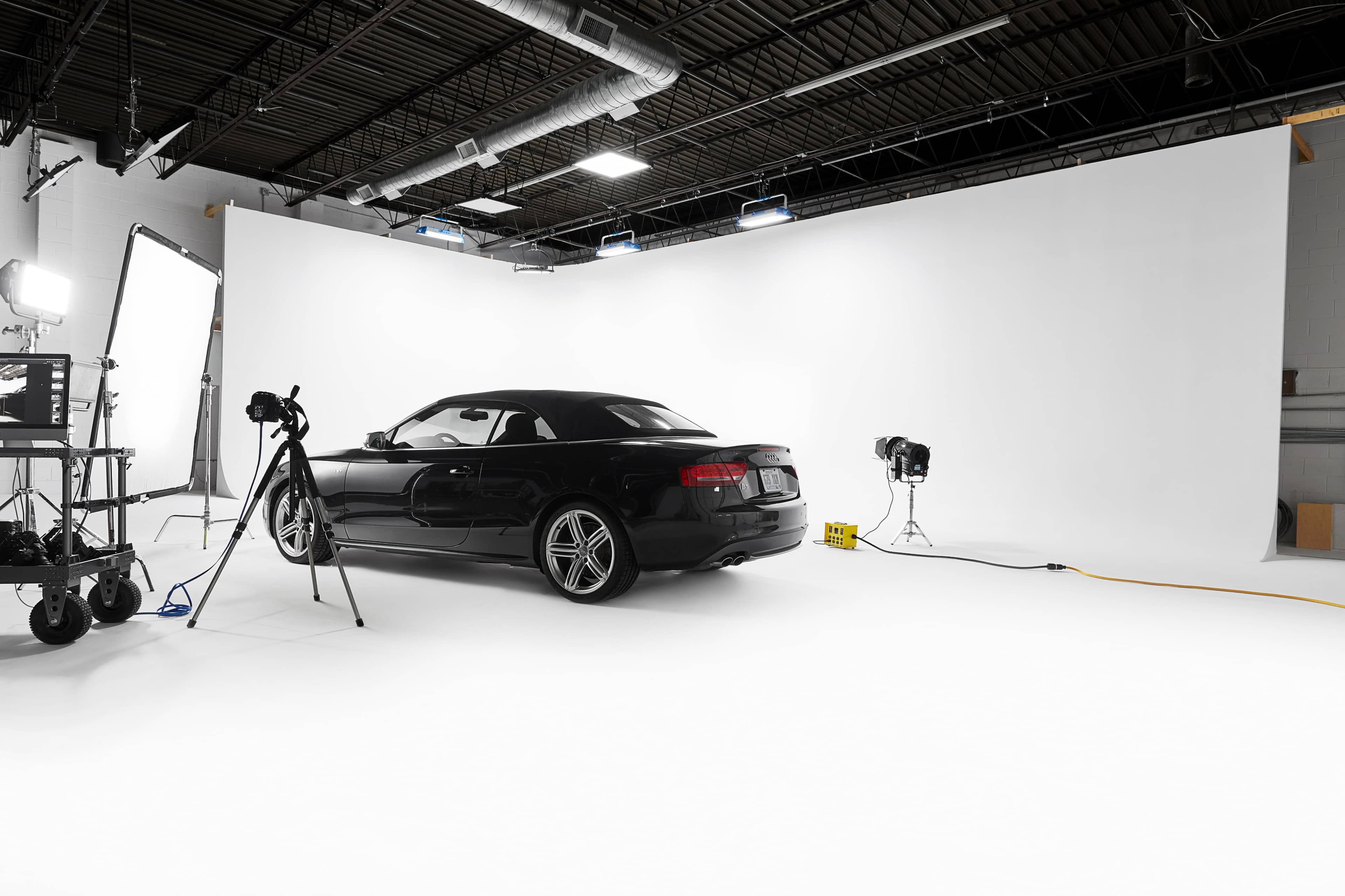 A black convertible car is positioned on a white backdrop in a photography studio equipped with lights and a camera setup.