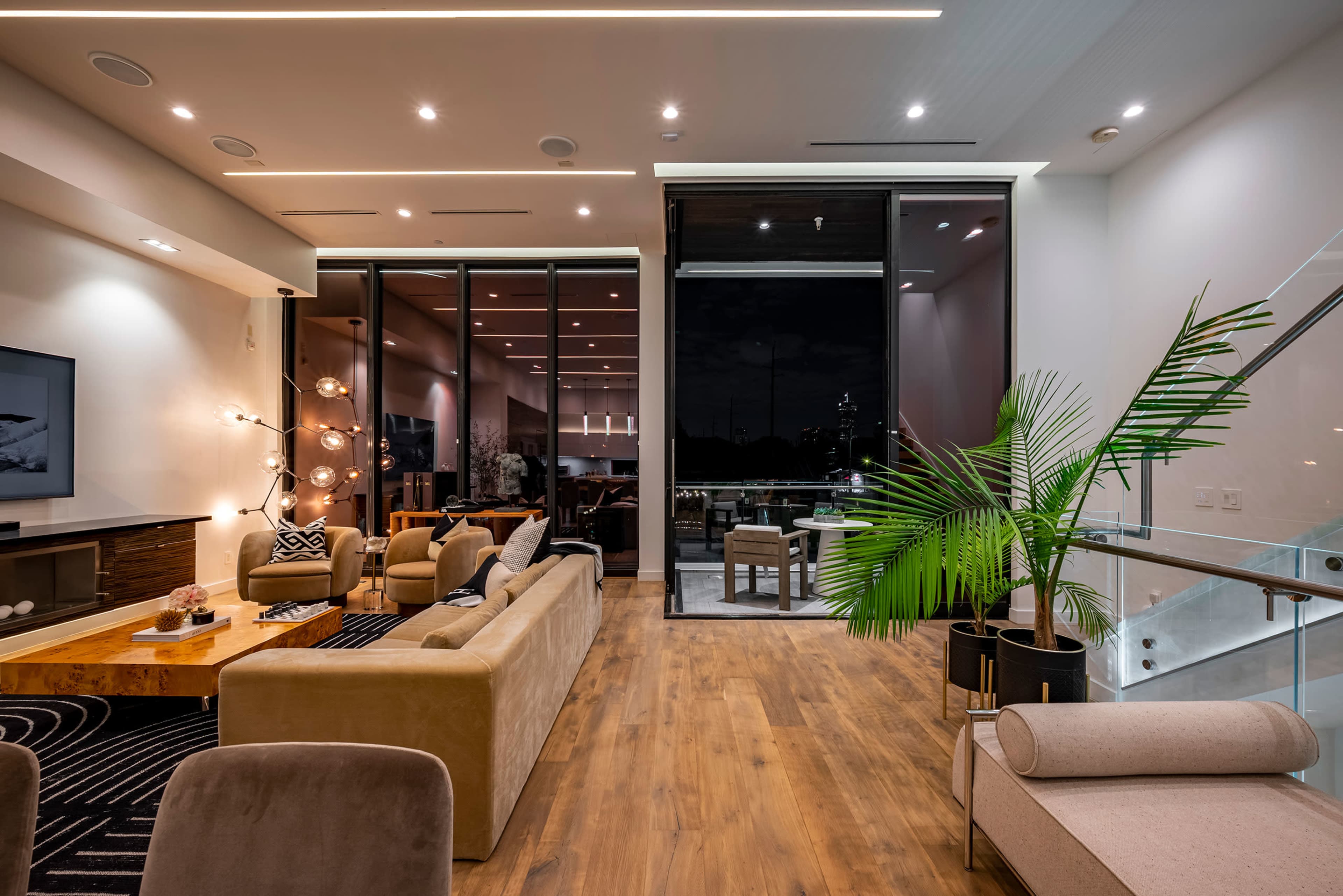 A modern living room with large windows, stylish furniture, and a decorative plant, all set against a backdrop of evening lighting.