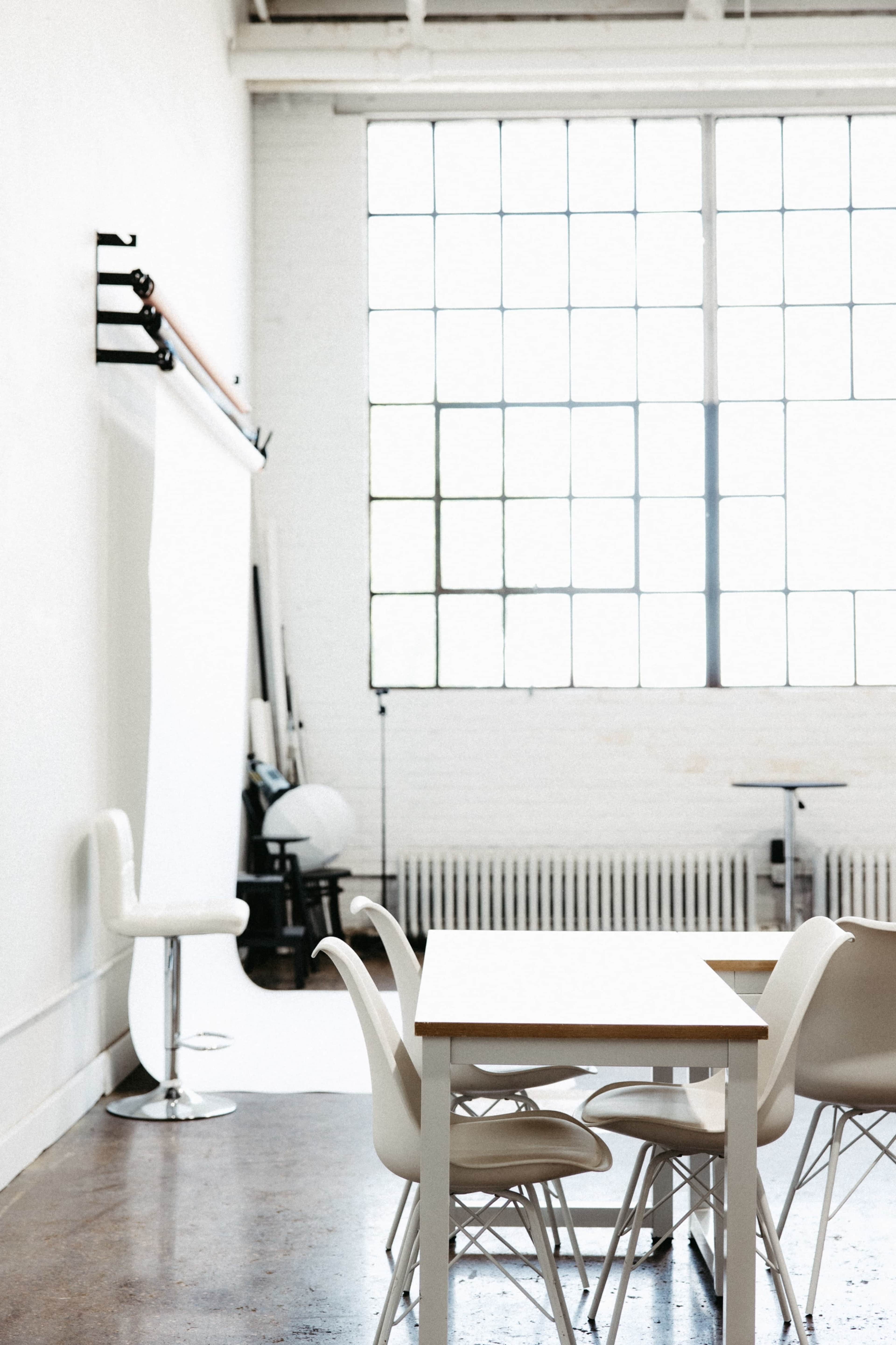 The image shows a minimalist studio space featuring a white backdrop, a table with chairs, and large windows providing natural light.