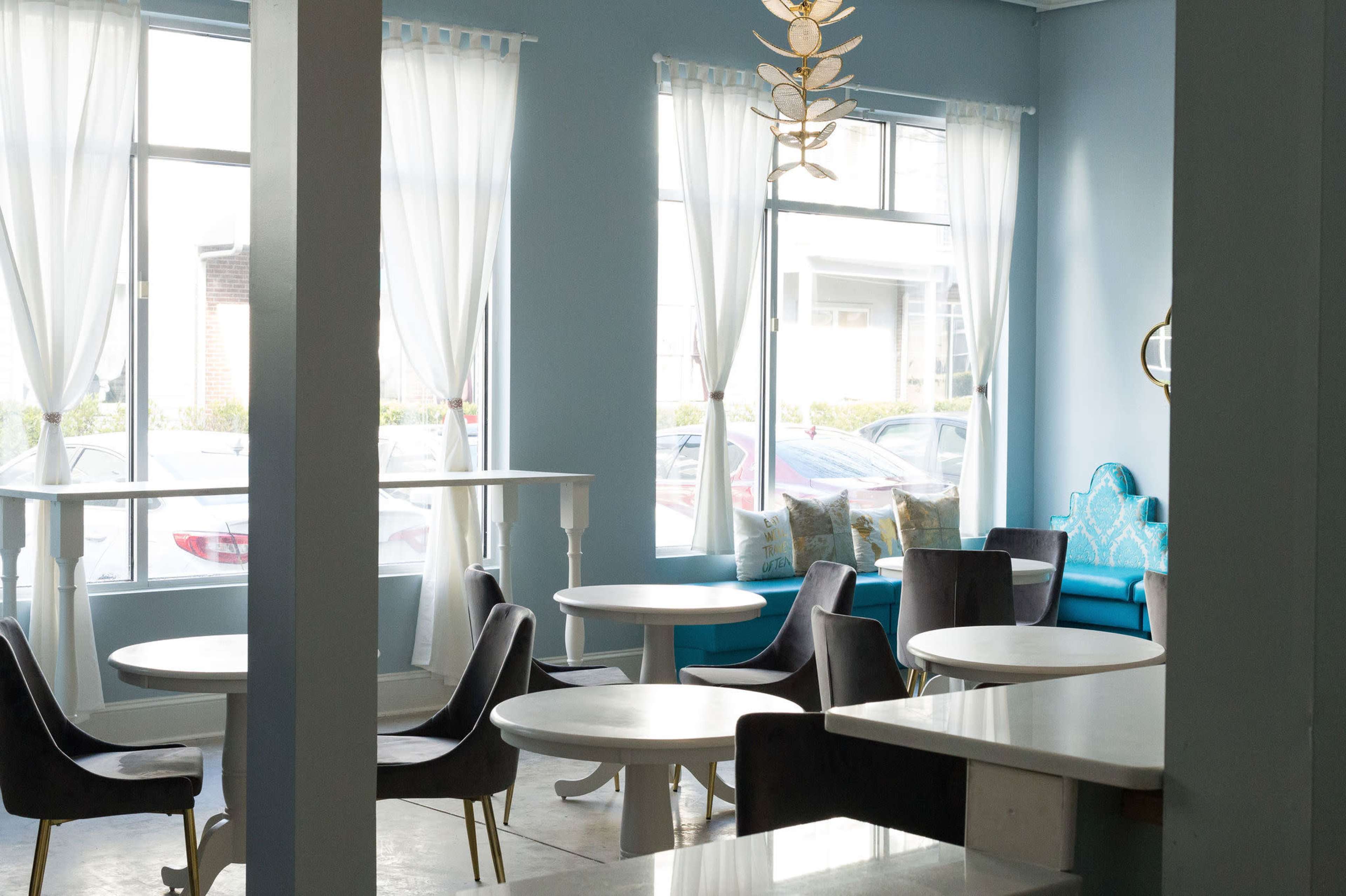 A brightly lit café interior with blue walls, large windows, and a mix of round tables and upholstered chairs.