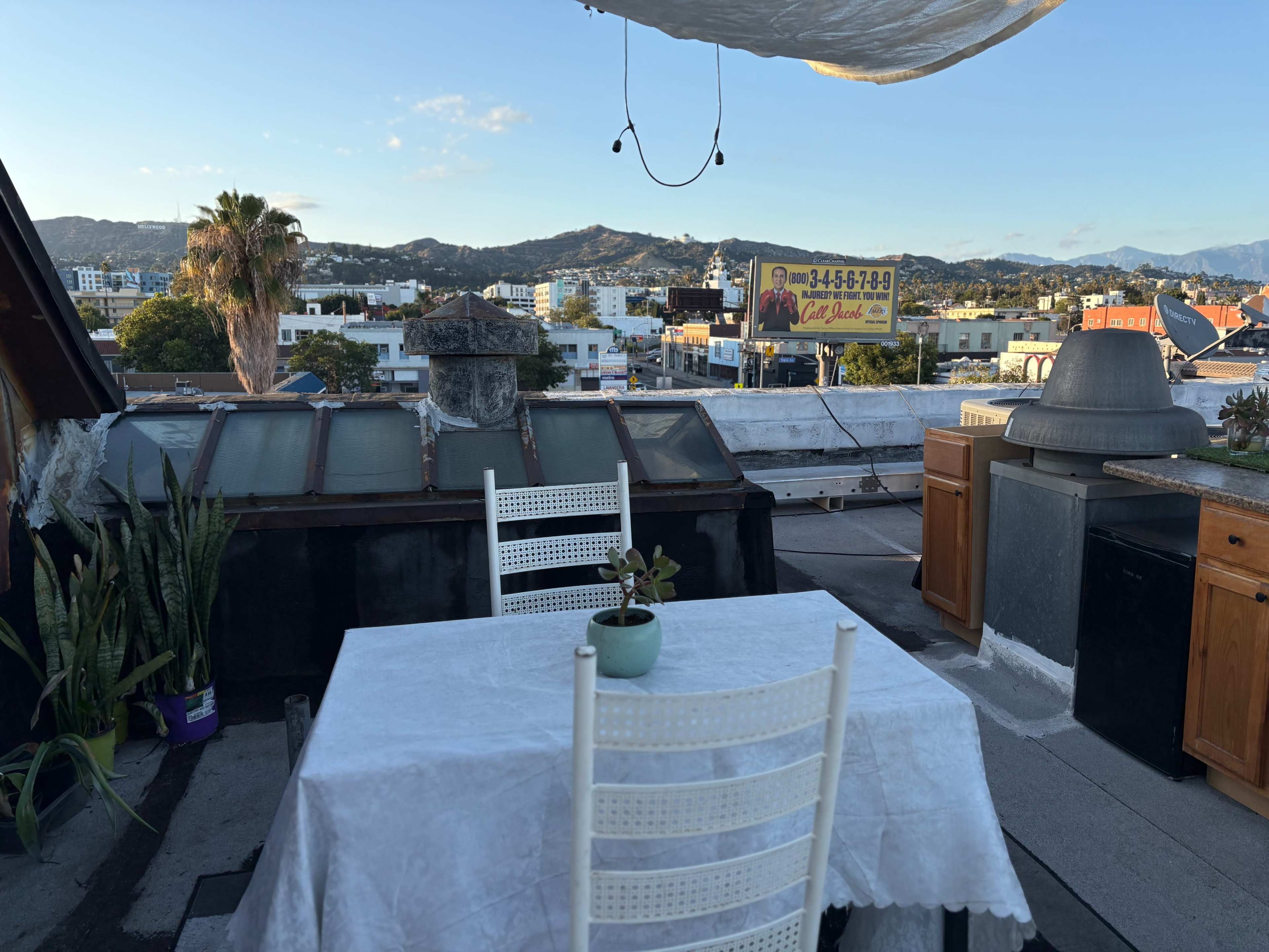 A white table with a small plant sits on a rooftop, surrounded by city views and mountains in the background.