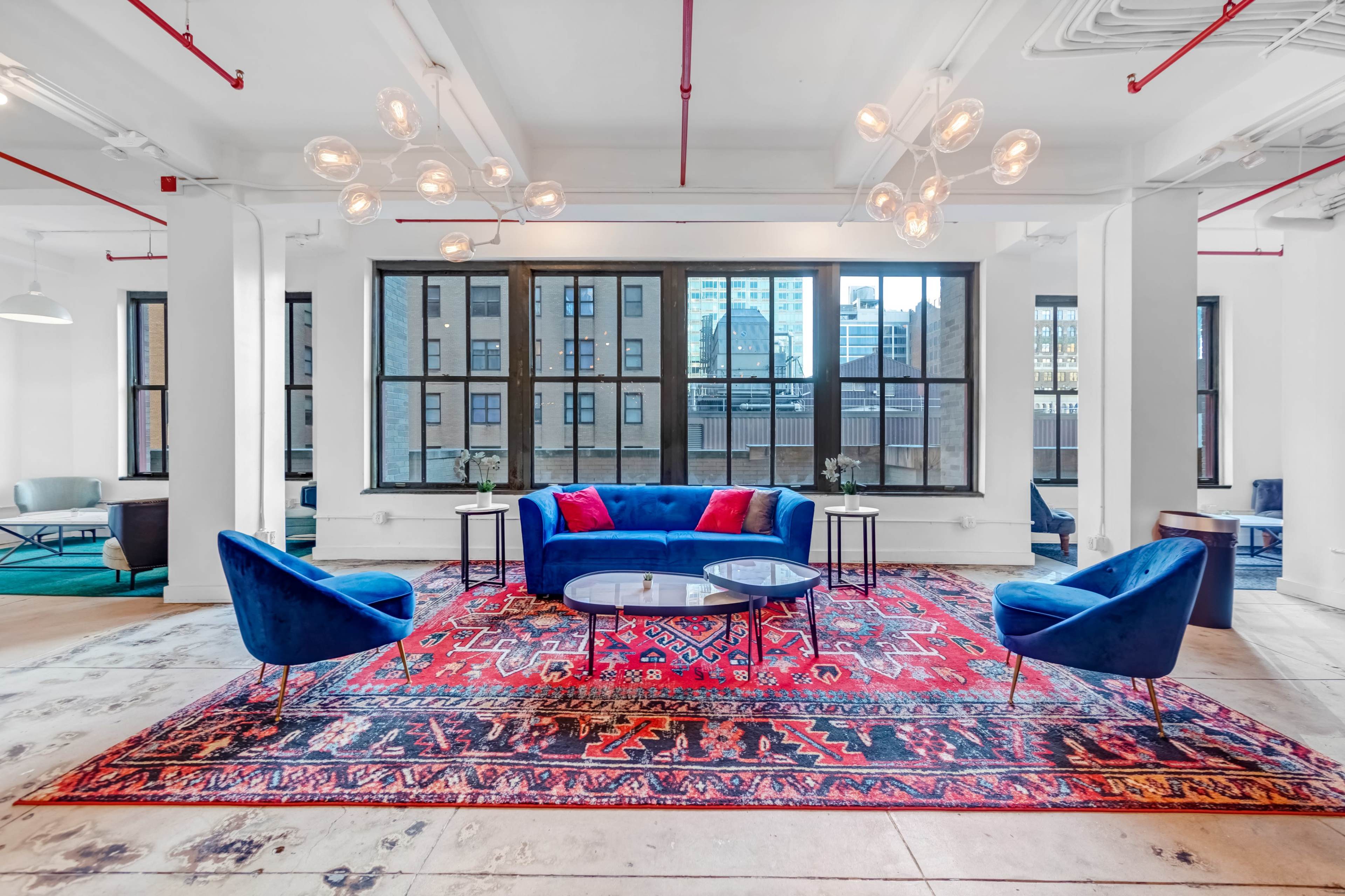 The image shows a modern, bright interior with a blue velvet sofa and matching armchairs arranged around a glass coffee table on a decorative red rug, with large windows providing a view of buildings outside.