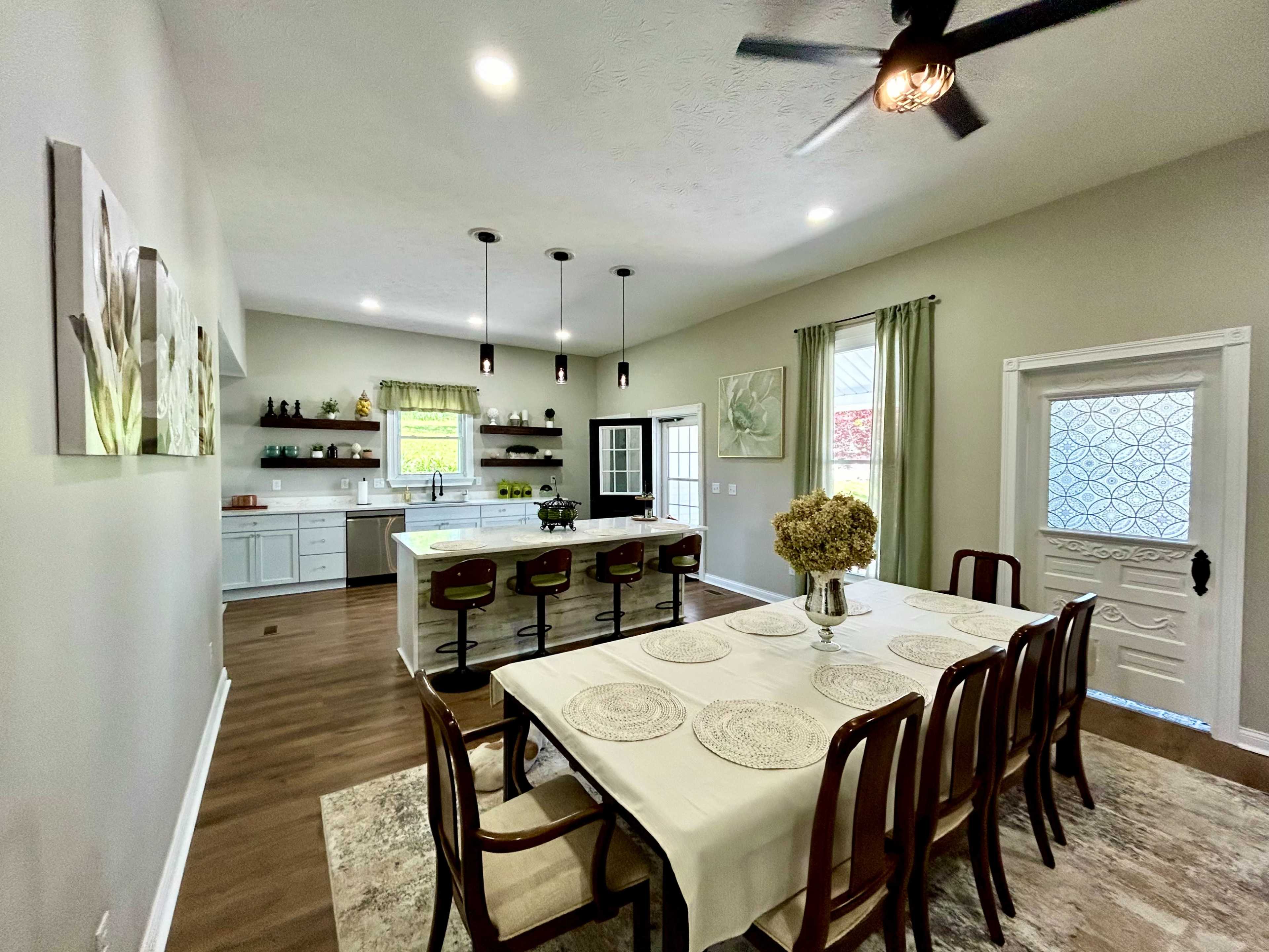 The image shows a modern kitchen and dining area featuring a long dining table, barstools at a kitchen counter, and large windows with natural light.