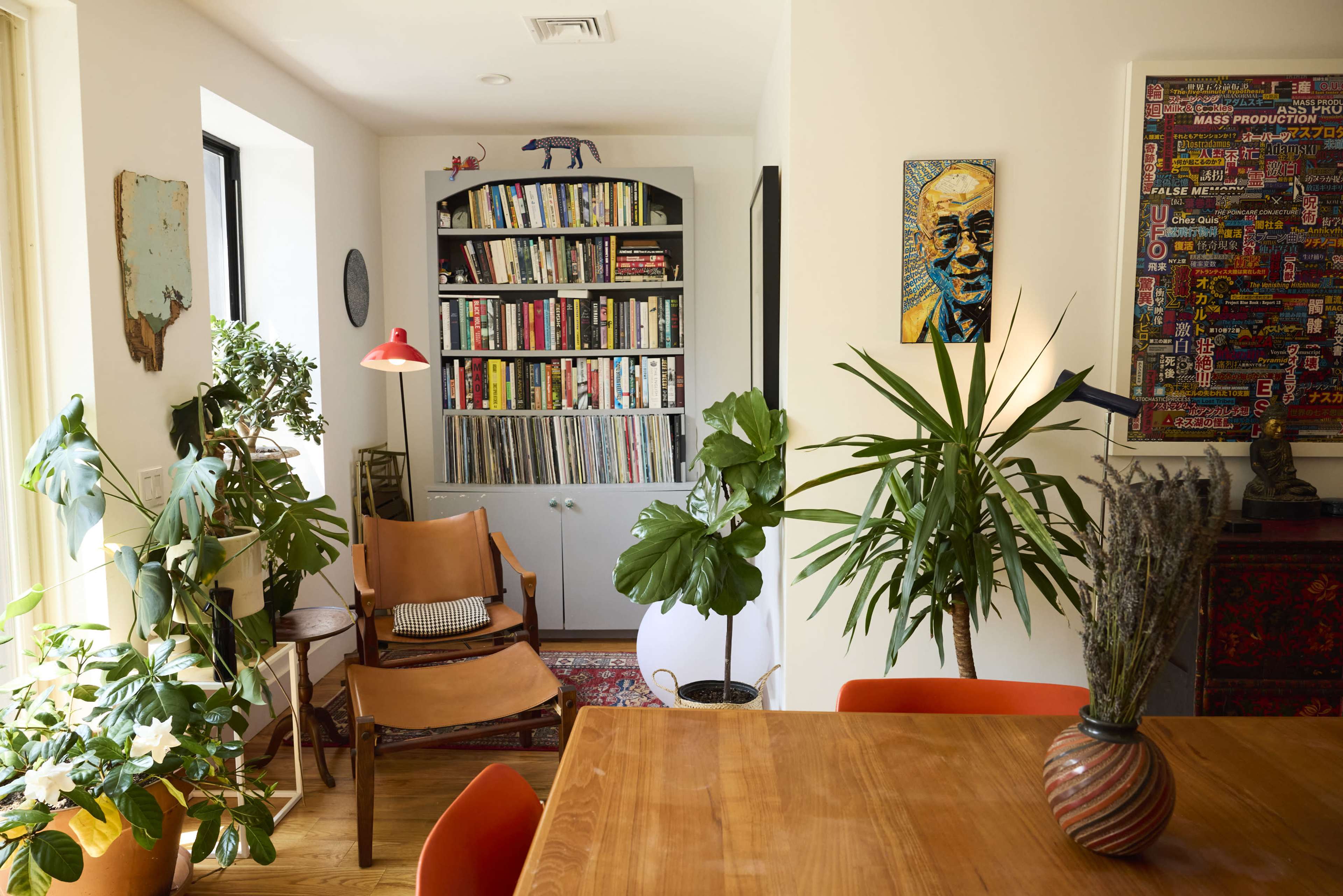 The image shows a cozy room with a wooden dining table, surrounded by plants, a bookshelf filled with books, and a mix of modern and vintage decor elements.