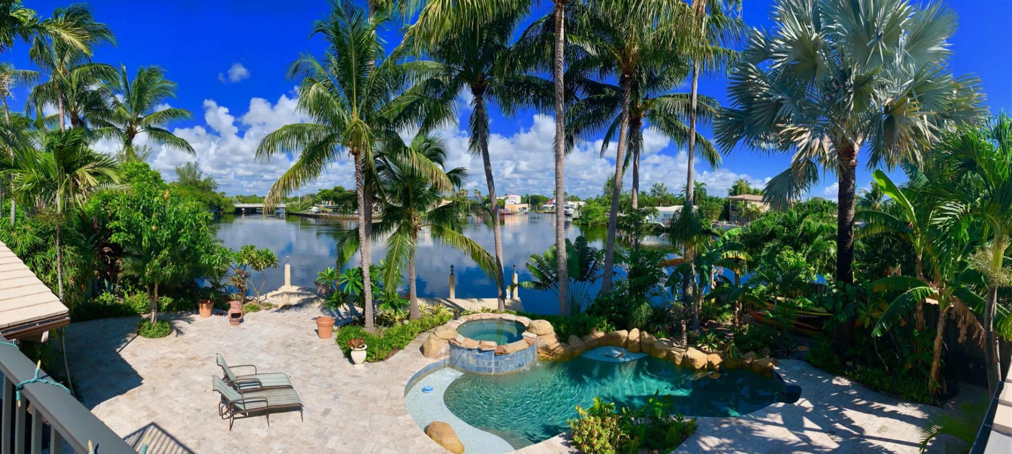 A landscaped backyard features a pool and spa area surrounded by palm trees and a waterfront view.
