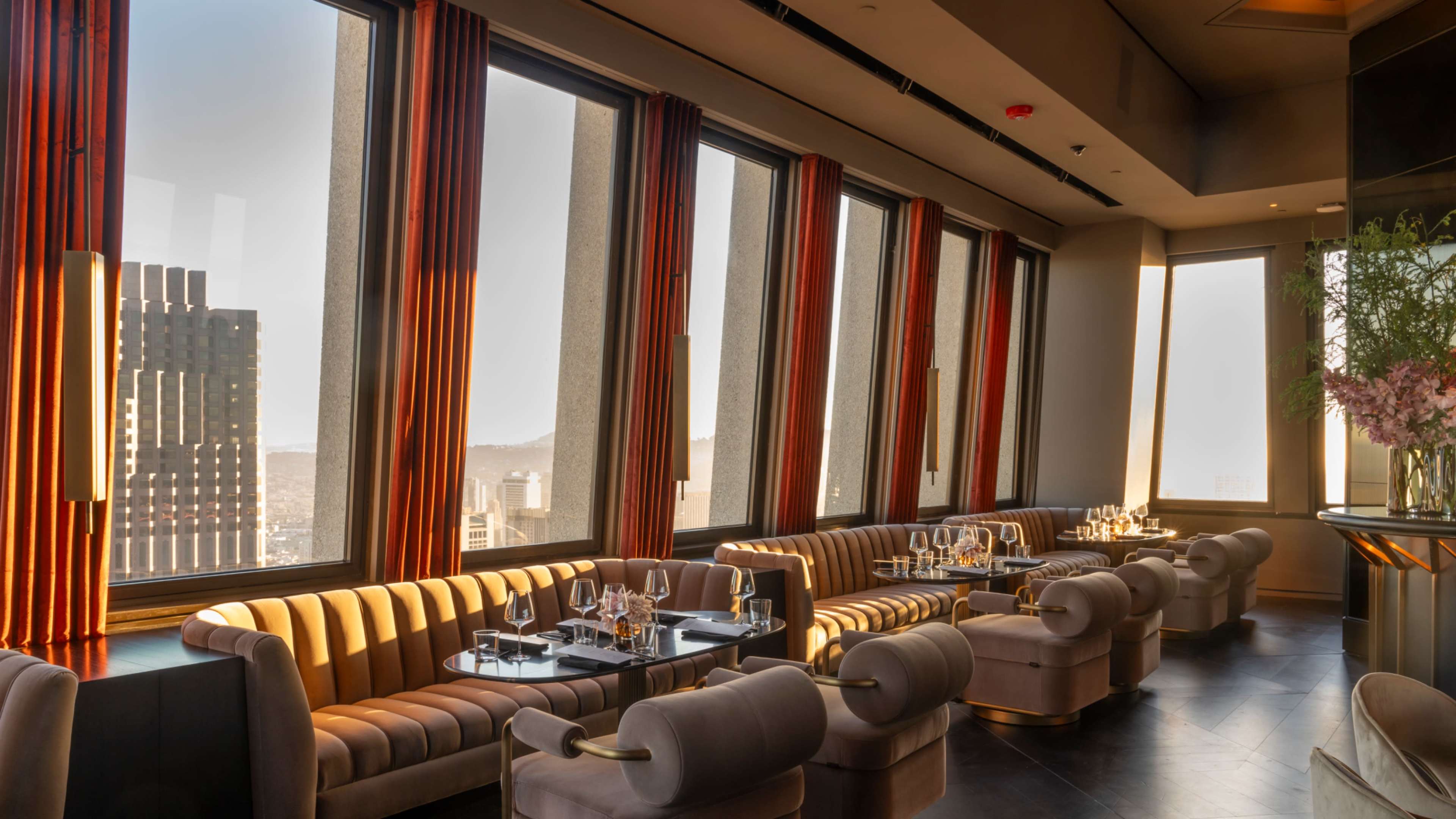 The image shows an upscale restaurant with plush seating, large windows offering a city view, and elegant table settings.
