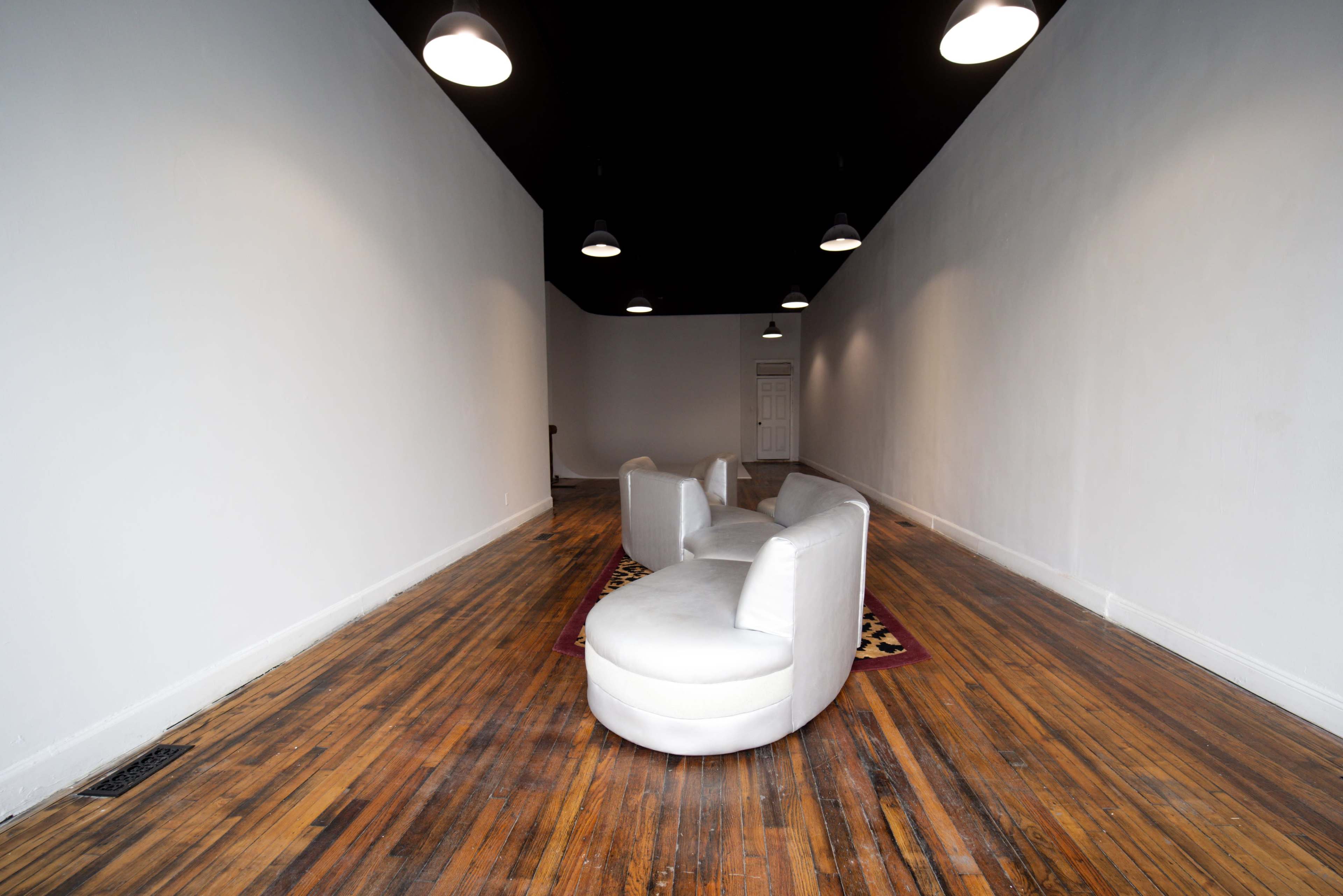 The image depicts a narrow, empty room with white walls and a black ceiling, featuring two white curved chairs on a patterned rug against a wooden floor.