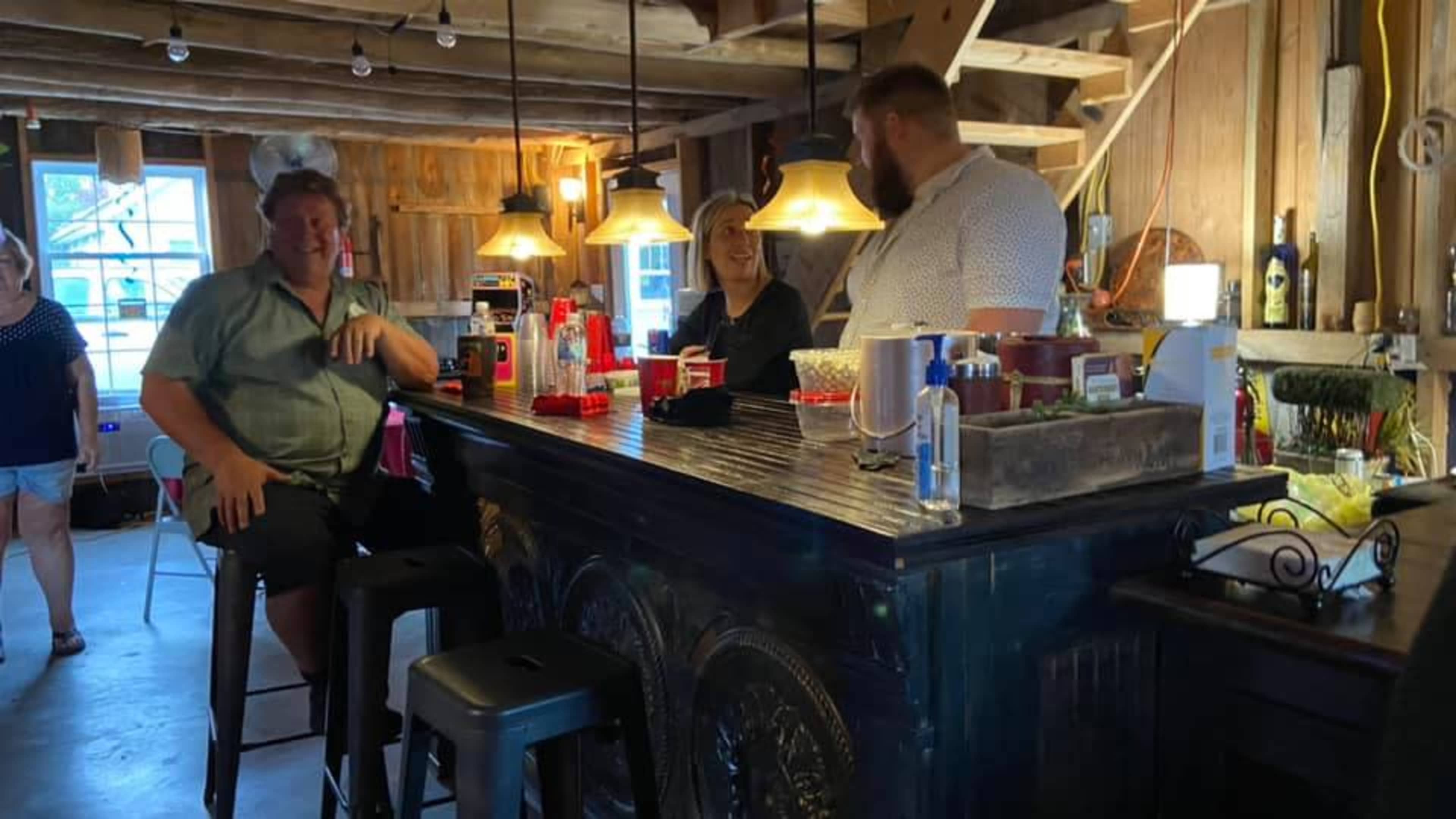 Three people are gathered at a bar in a wooden interior while others stand in the background.