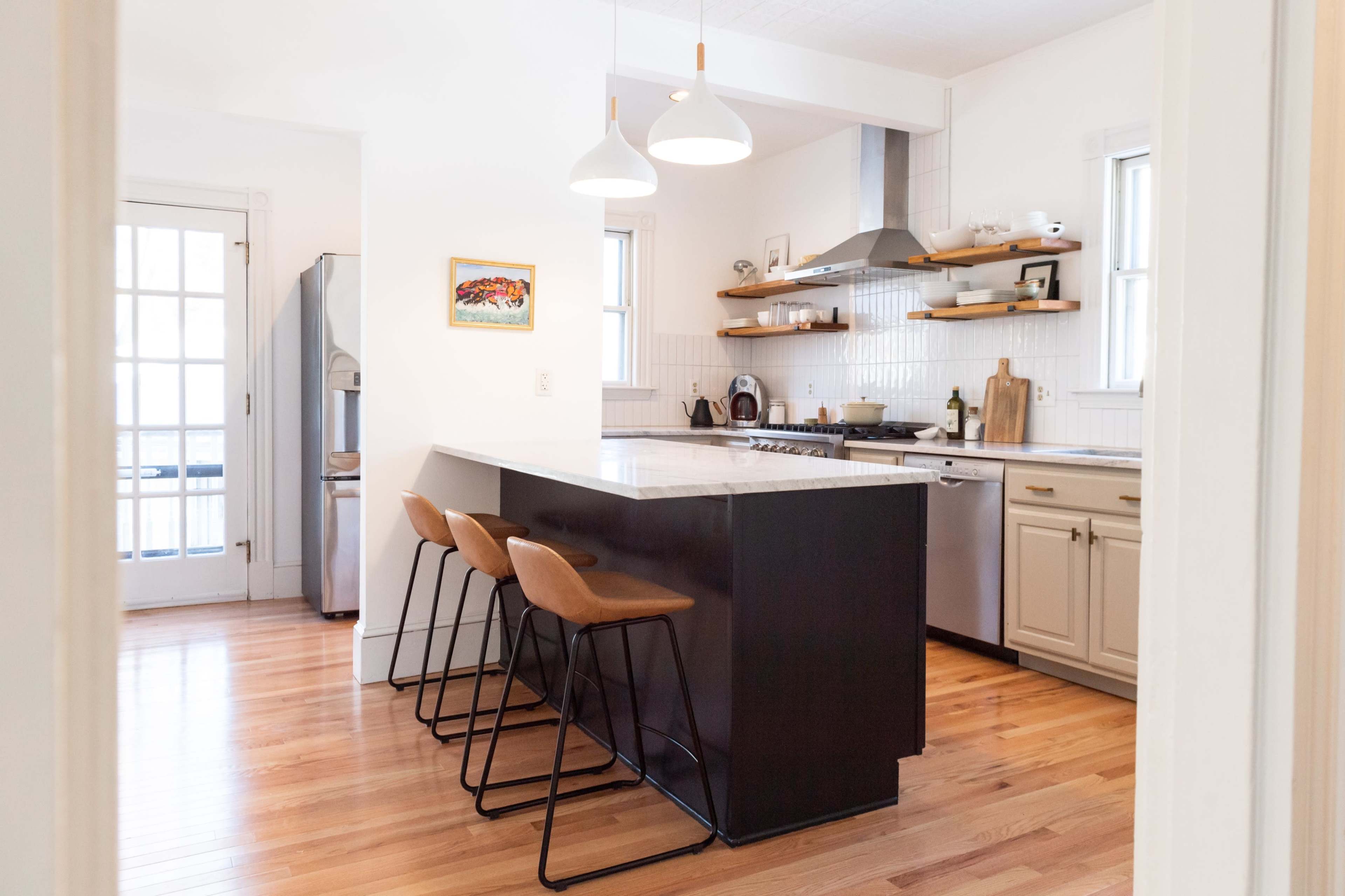 A modern kitchen with a central island and three bar stools, featuring white cabinetry, stainless steel appliances, and wooden floors.