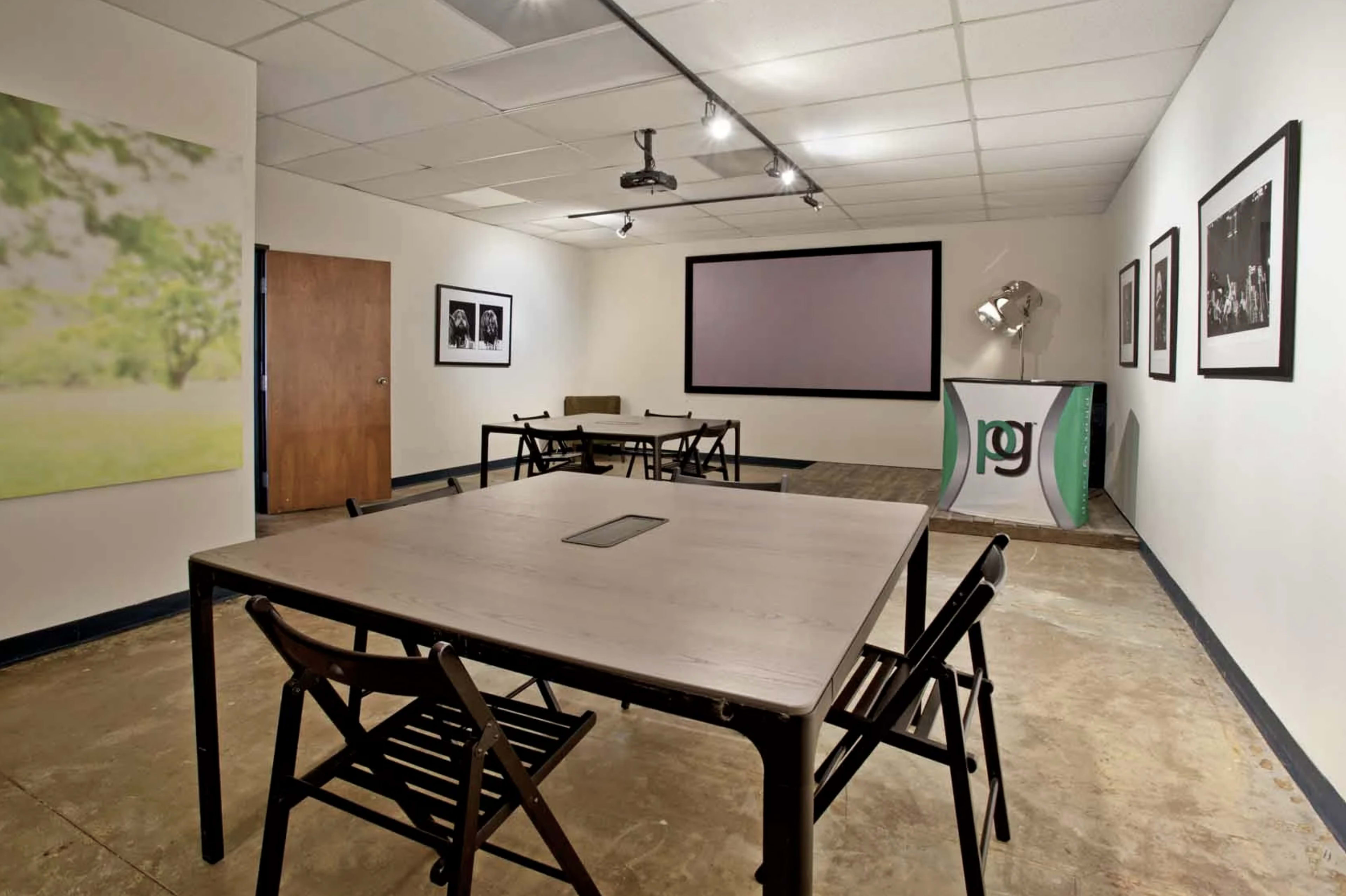 A spacious meeting room with two tables, several chairs, a projector screen, and framed photographs on the walls.