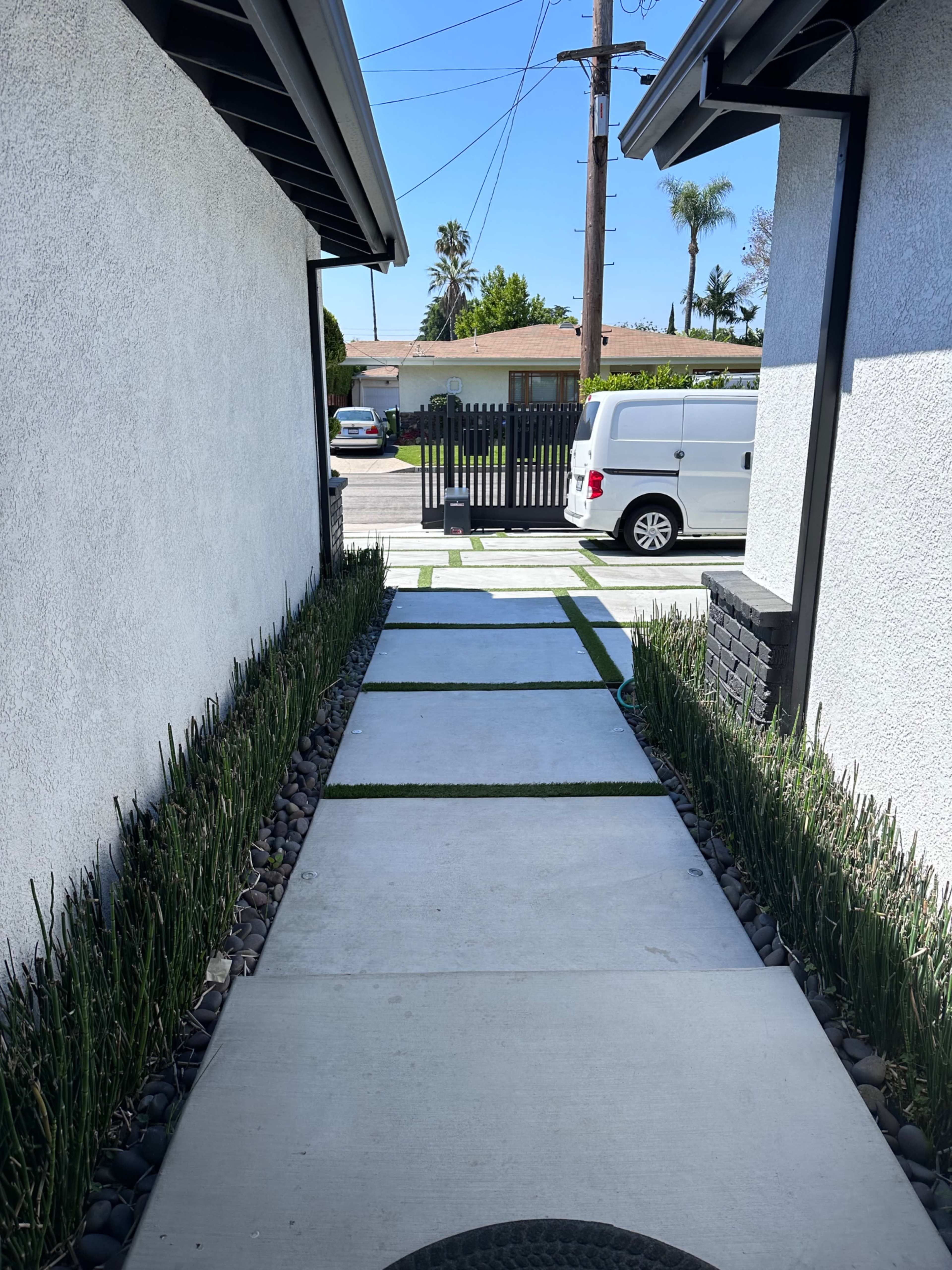 A walkway lined with grass and decorative stones, leading between two white buildings to a street with parked vehicles and a fence in the background.