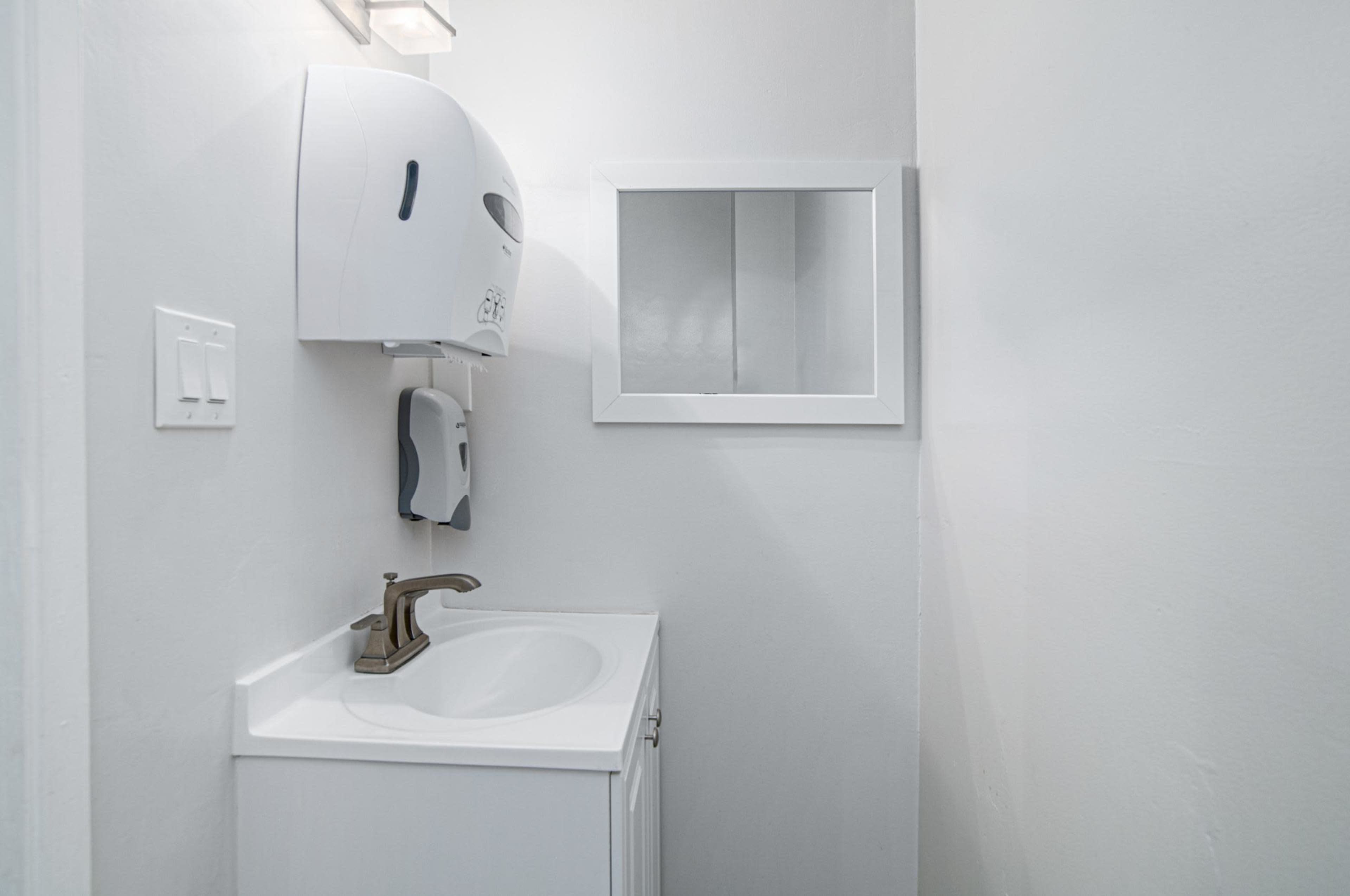 A small bathroom features a sink, a wall-mounted paper towel dispenser, and a mirror against a plain white wall.