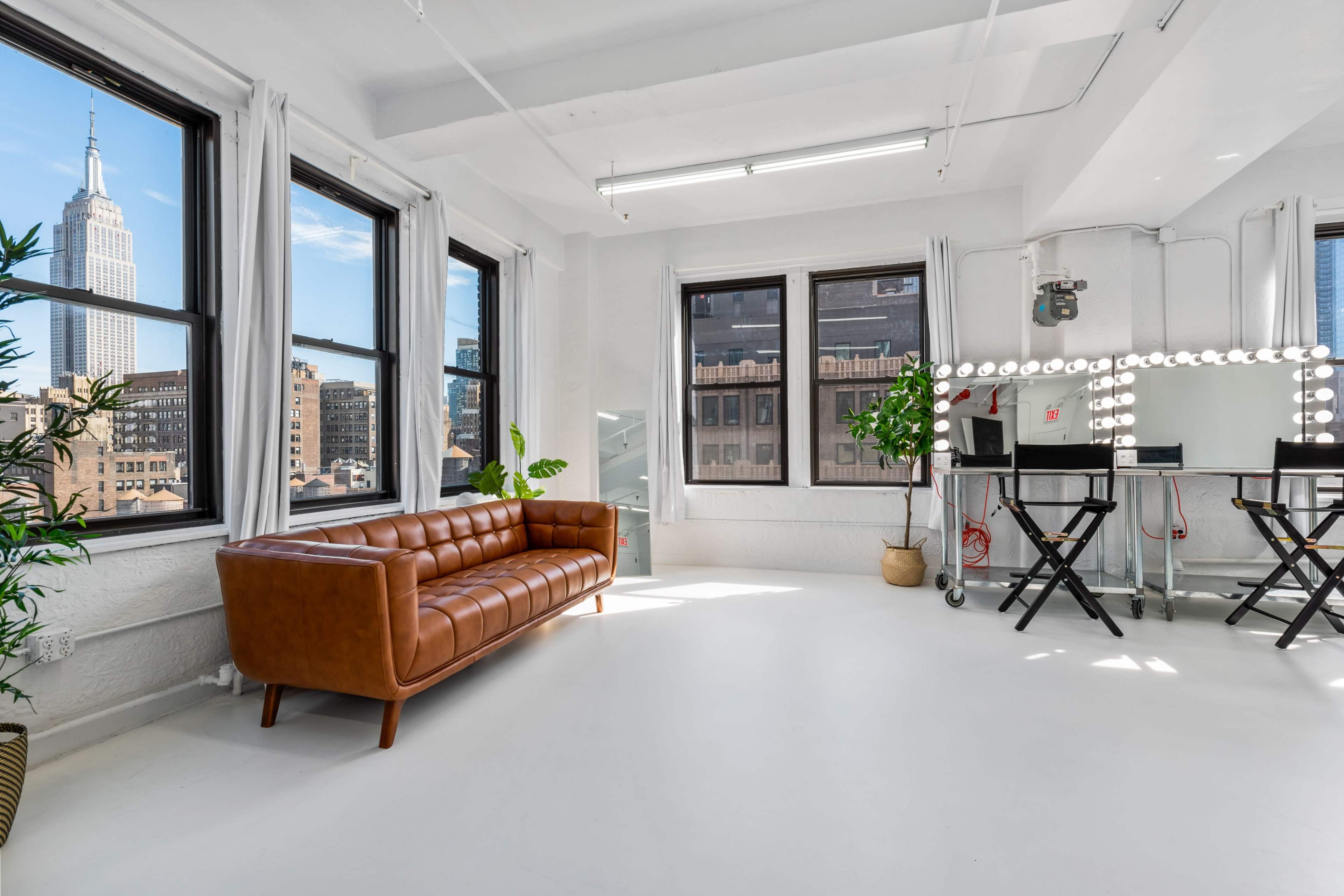 A bright, minimalist studio features a brown leather couch, a vanity with lights, and large windows showcasing the Empire State Building in the background.