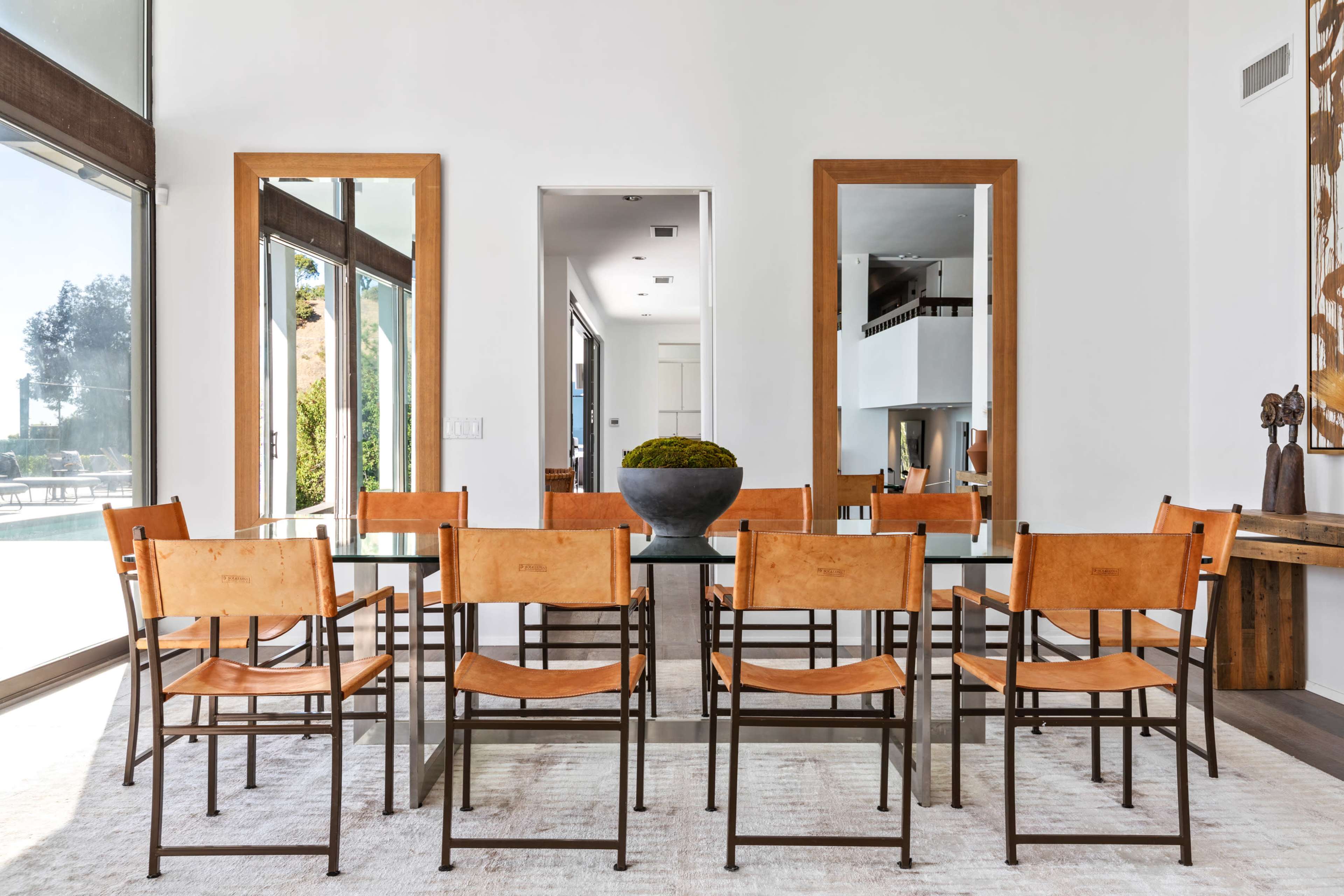 A modern dining room features a glass table surrounded by eight brown leather chairs, with large mirrors and windows reflecting natural light.