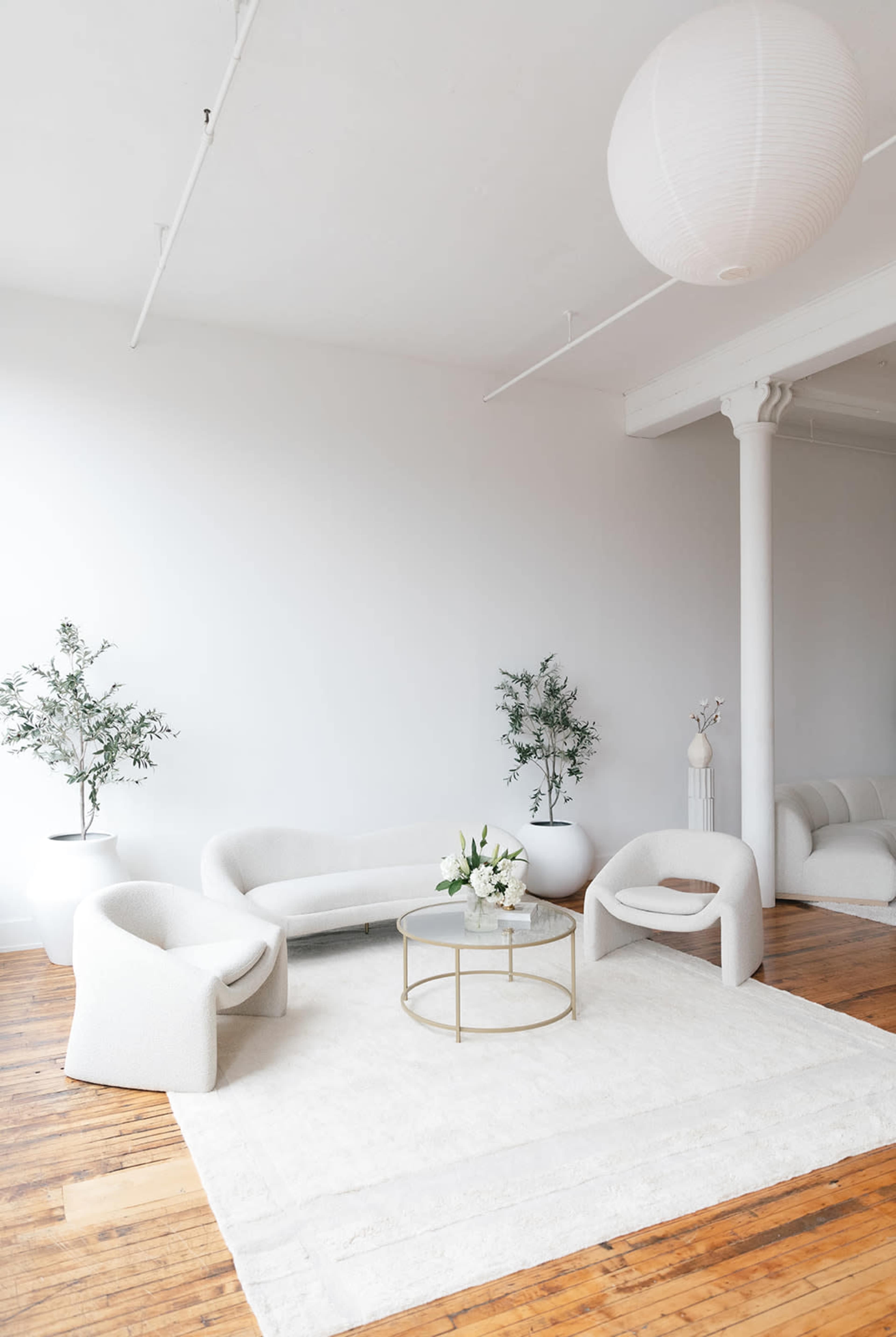A modern living room with a minimalist design featuring a white sofa, two armchairs, a round coffee table with flowers, and wooden flooring.