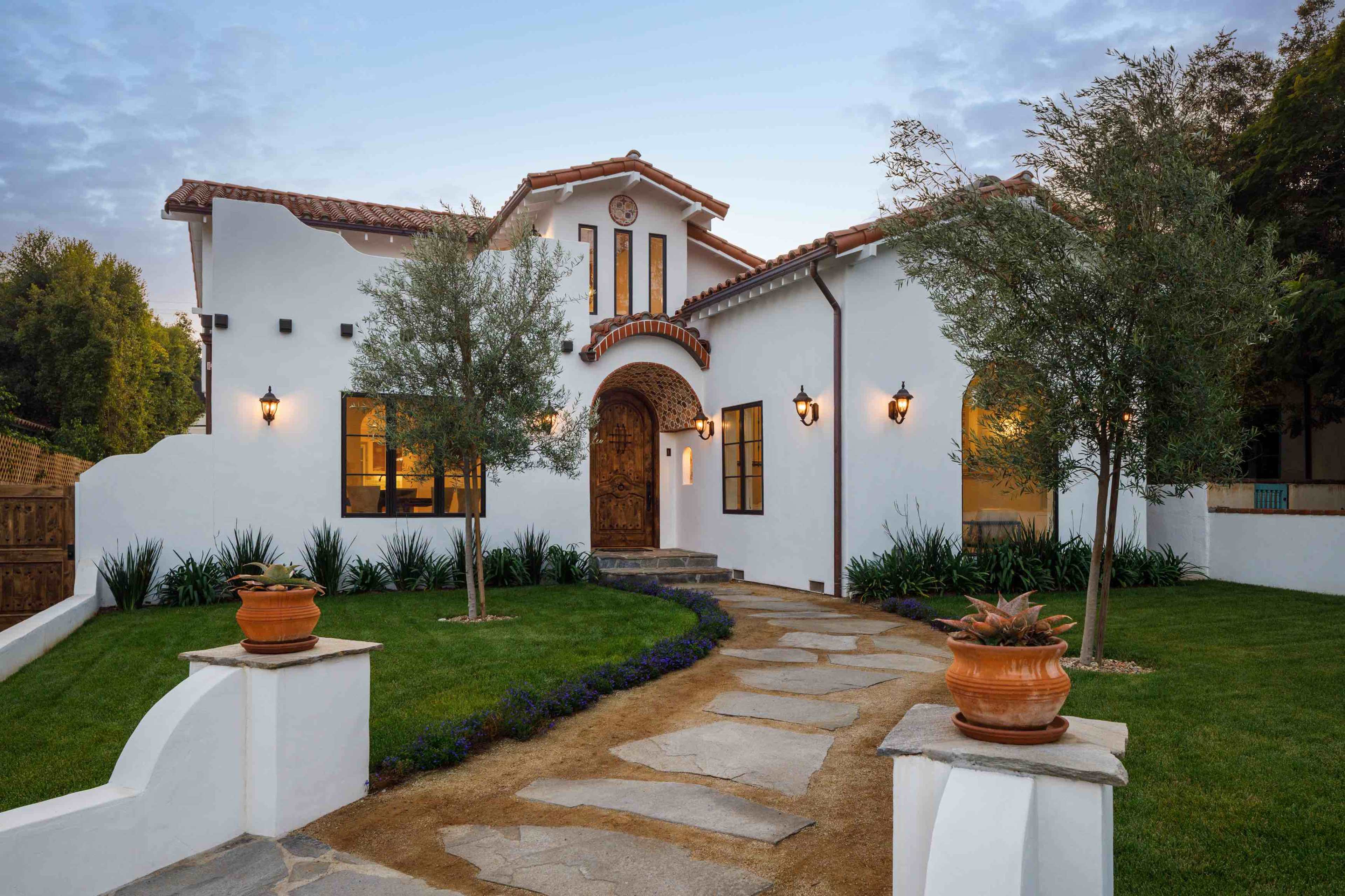 A white stucco house with a tile roof, surrounded by manicured landscaping and a stone walkway leading to the front entrance.