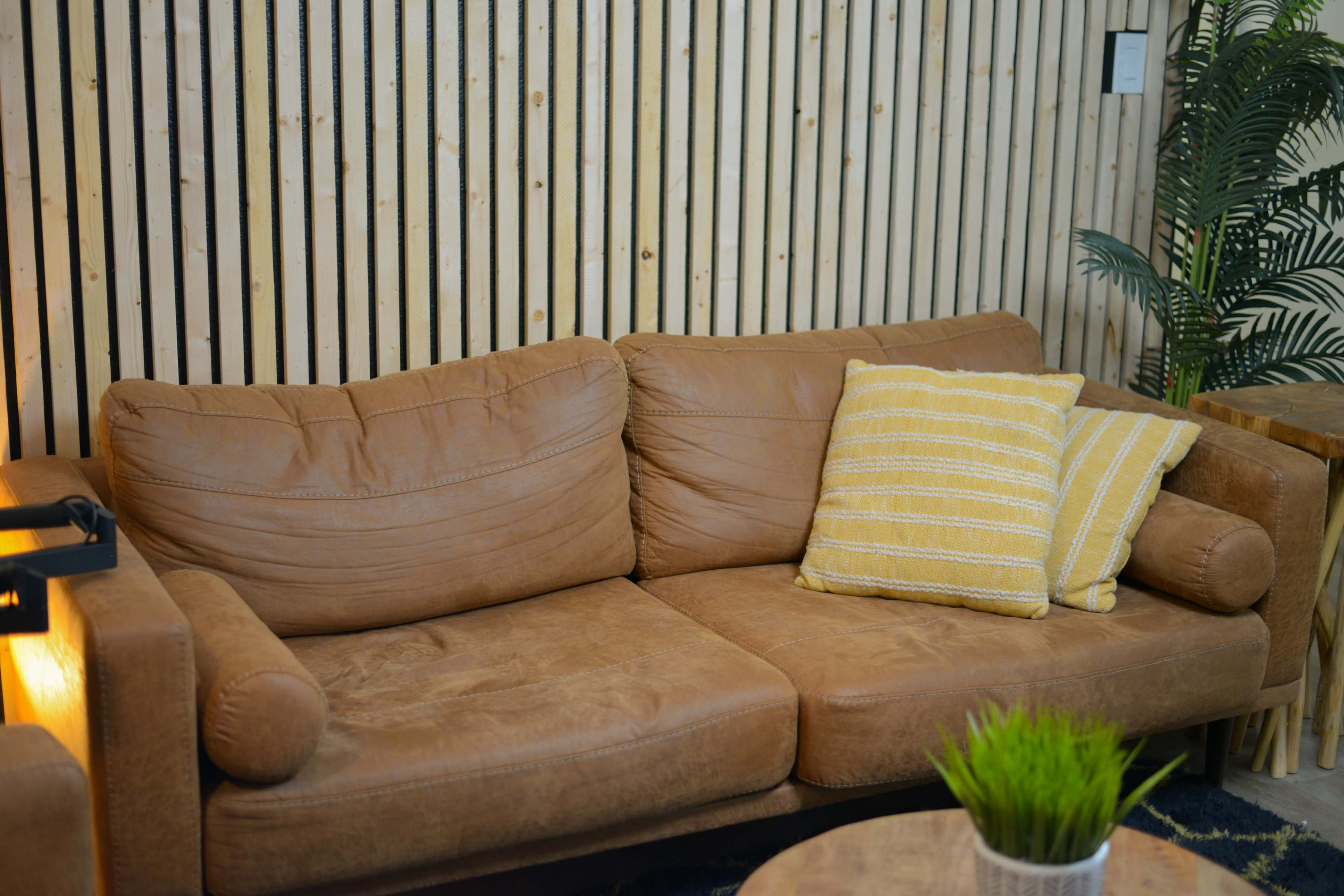 A brown leather couch with two pillows, situated in a room with wooden slat walls and a small plant nearby.