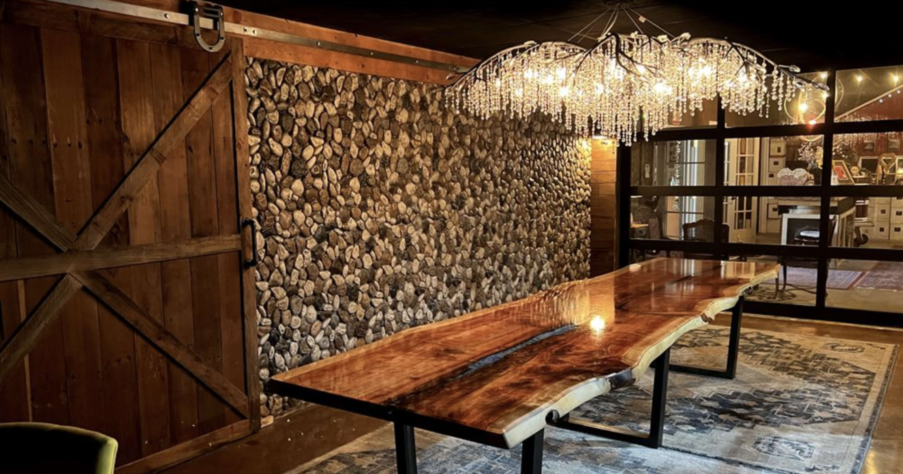 A rustic dining area featuring a large wooden table with a live edge, set against a stone accent wall and complemented by a contemporary chandelier above.