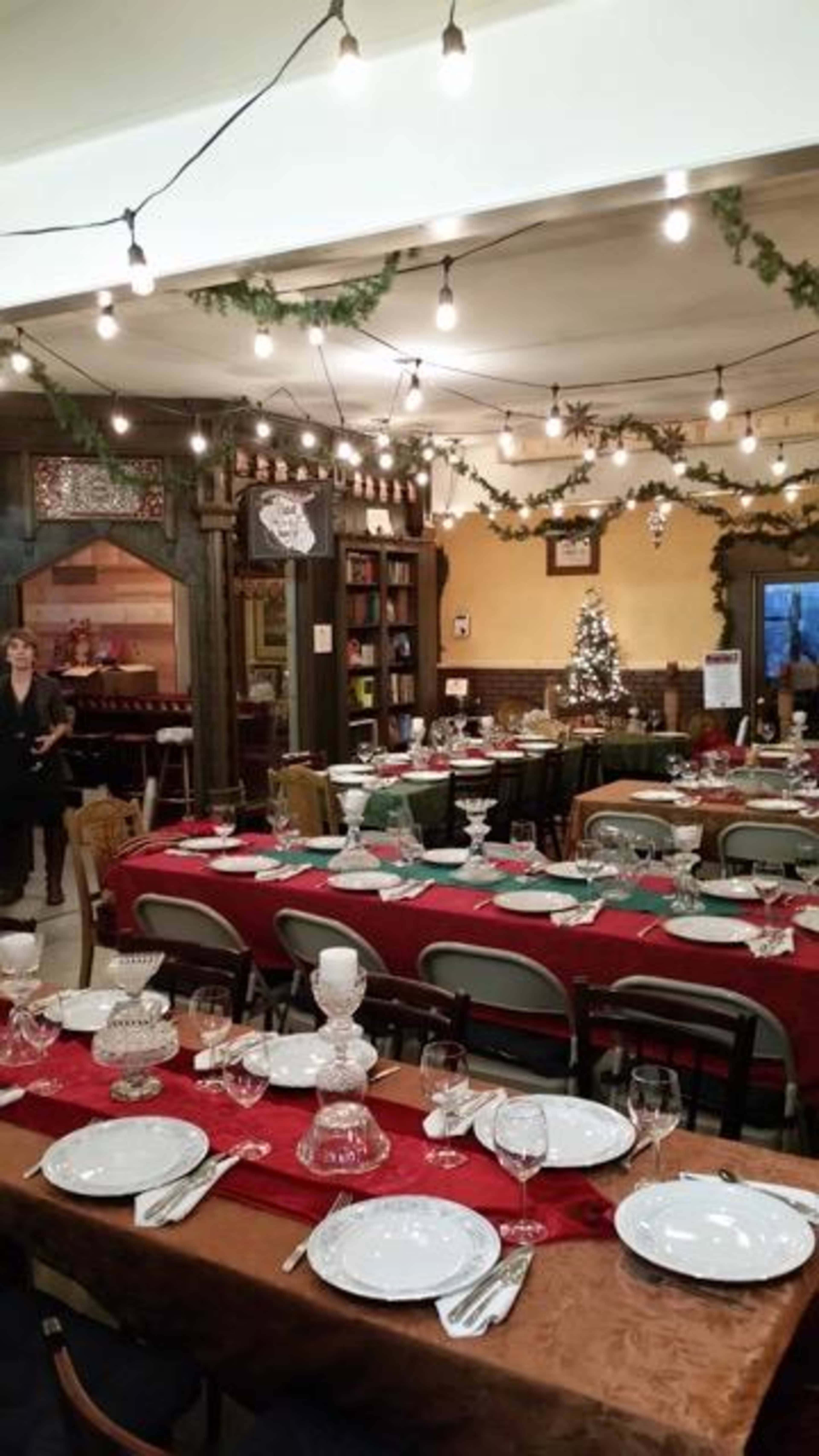 A dining area decorated for a festive gathering, featuring multiple tables set with plates, glasses, and tablecloths, and adorned with string lights and holiday decorations.
