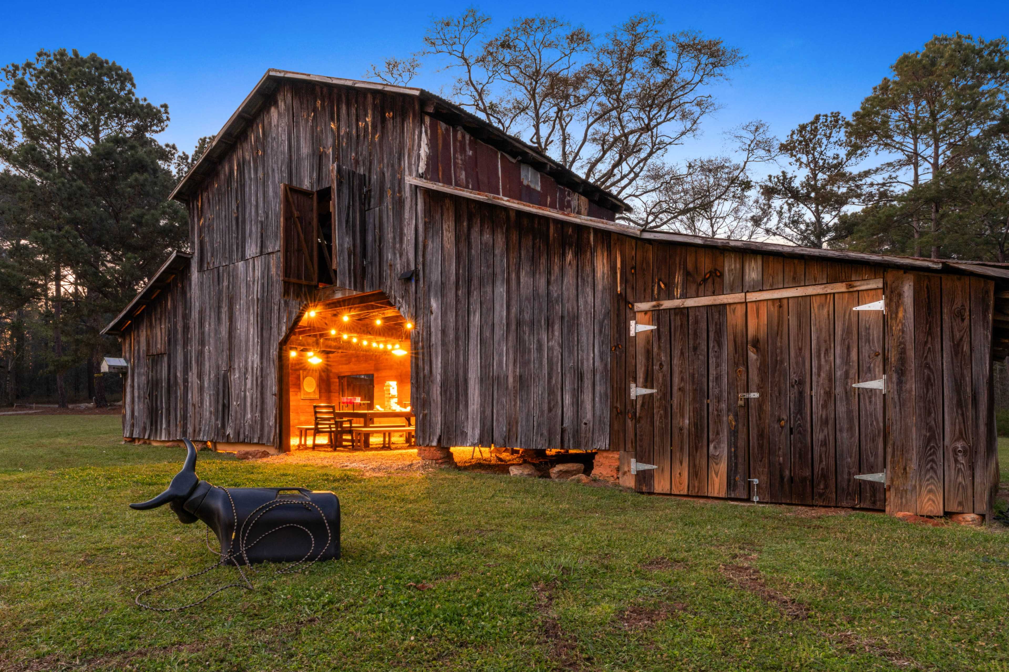 A rustic wooden barn with a partially open door reveals a warm, inviting interior adorned with string lights, while a mechanical bull sits outside on the grass.