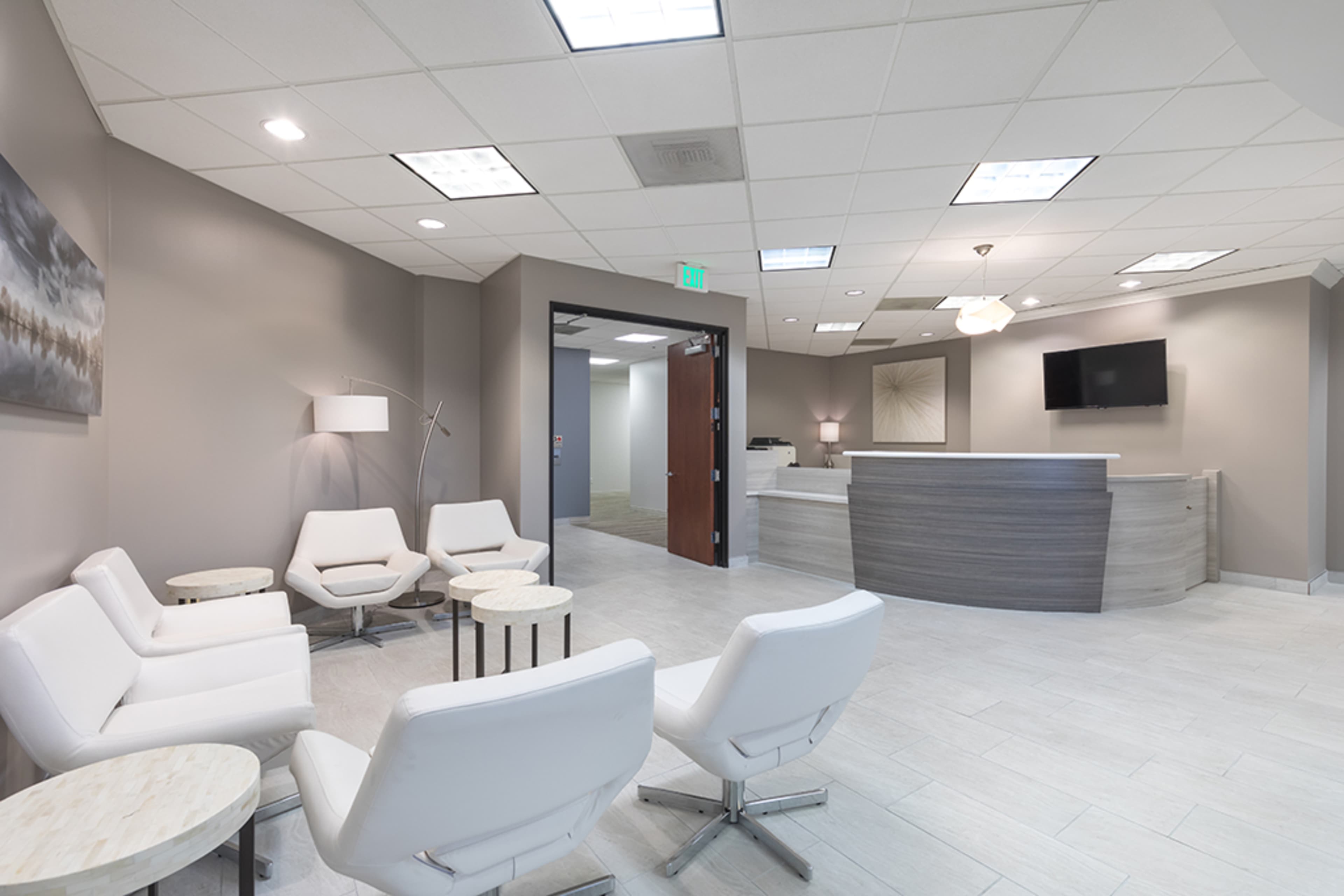A modern waiting room features several white chairs and small tables, with a reception desk in the background and a flat-screen TV mounted on the wall.