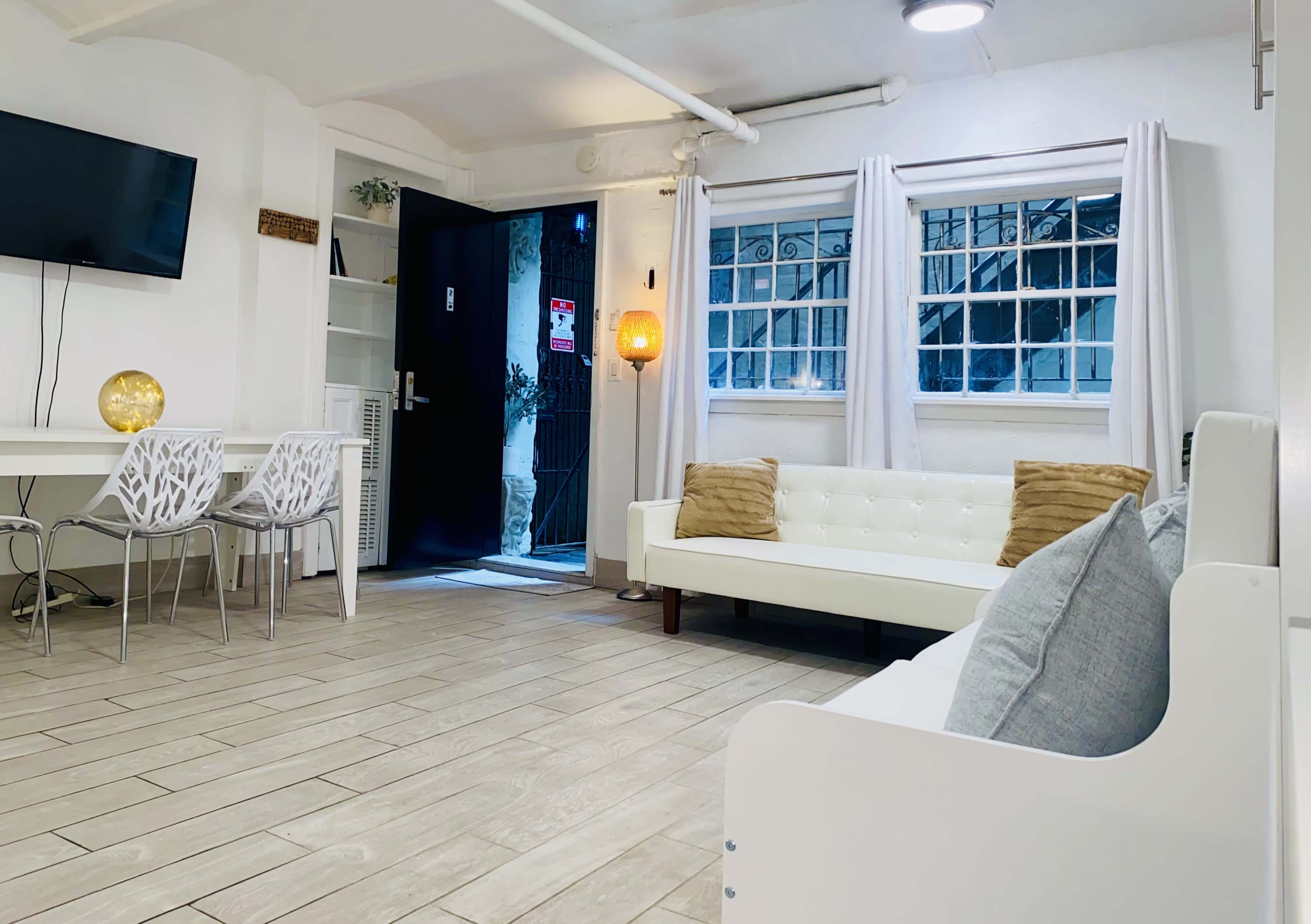 A bright, minimalist living space featuring a white sofa, a dining area with four chairs, and large windows with light curtains.